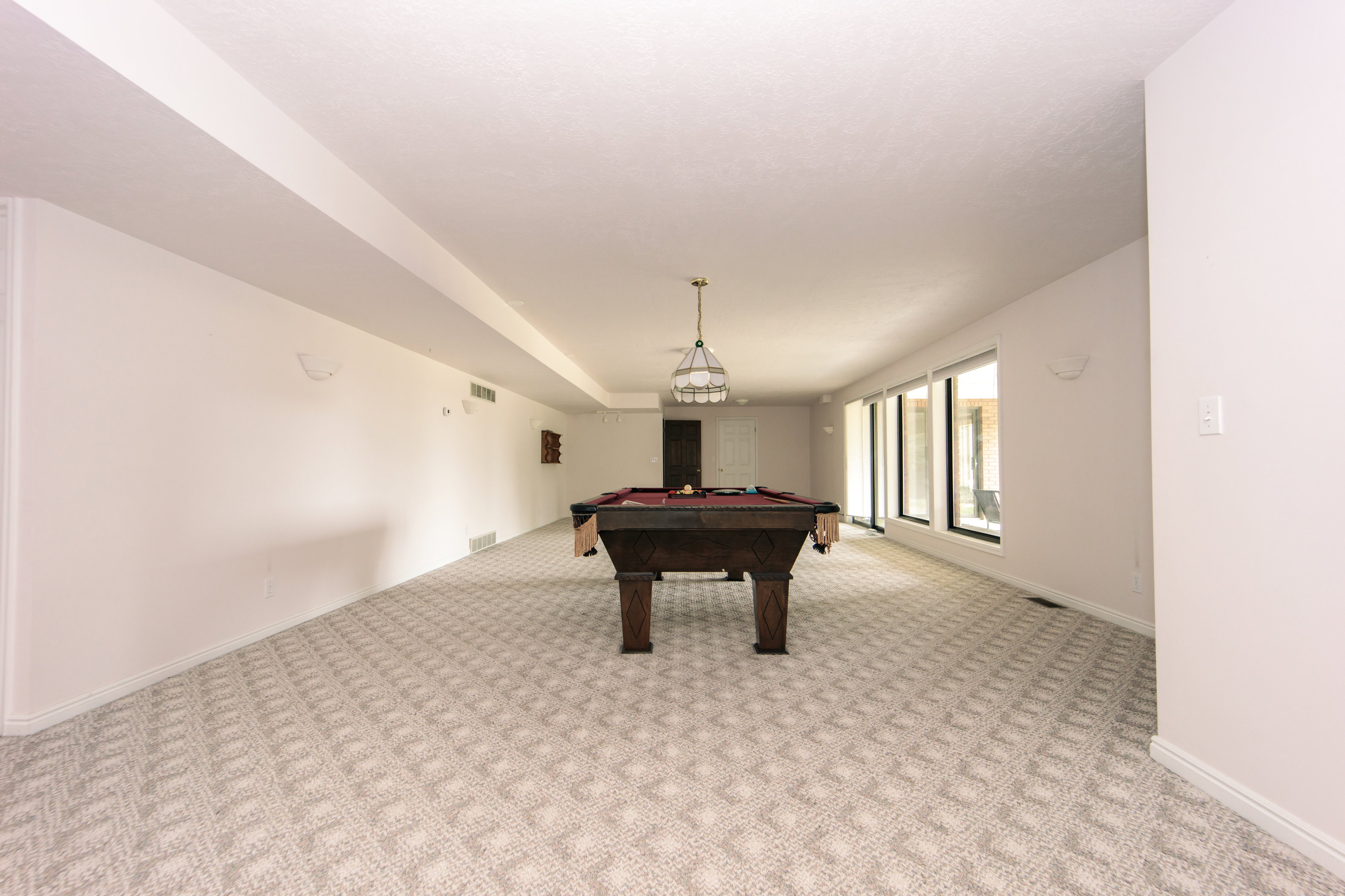 Spacious entertainment area featuring patterned carpeting, recessed ceiling beams, a central chandelier, and multiple large windows providing ample natural light