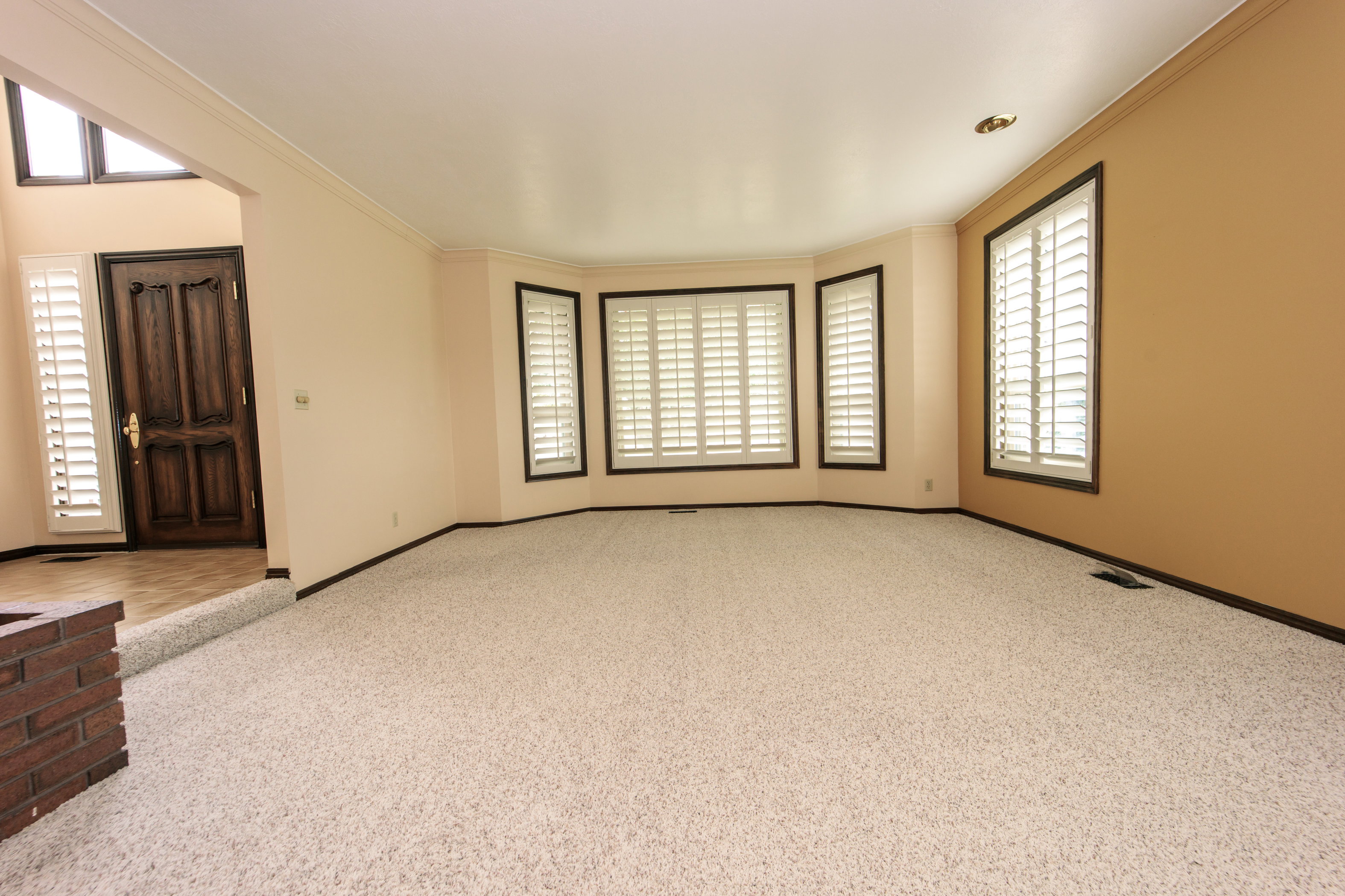 Spacious living area featuring light-colored carpeting, a bay window with plantation shutters, crown molding, and a brick-faced half-wall