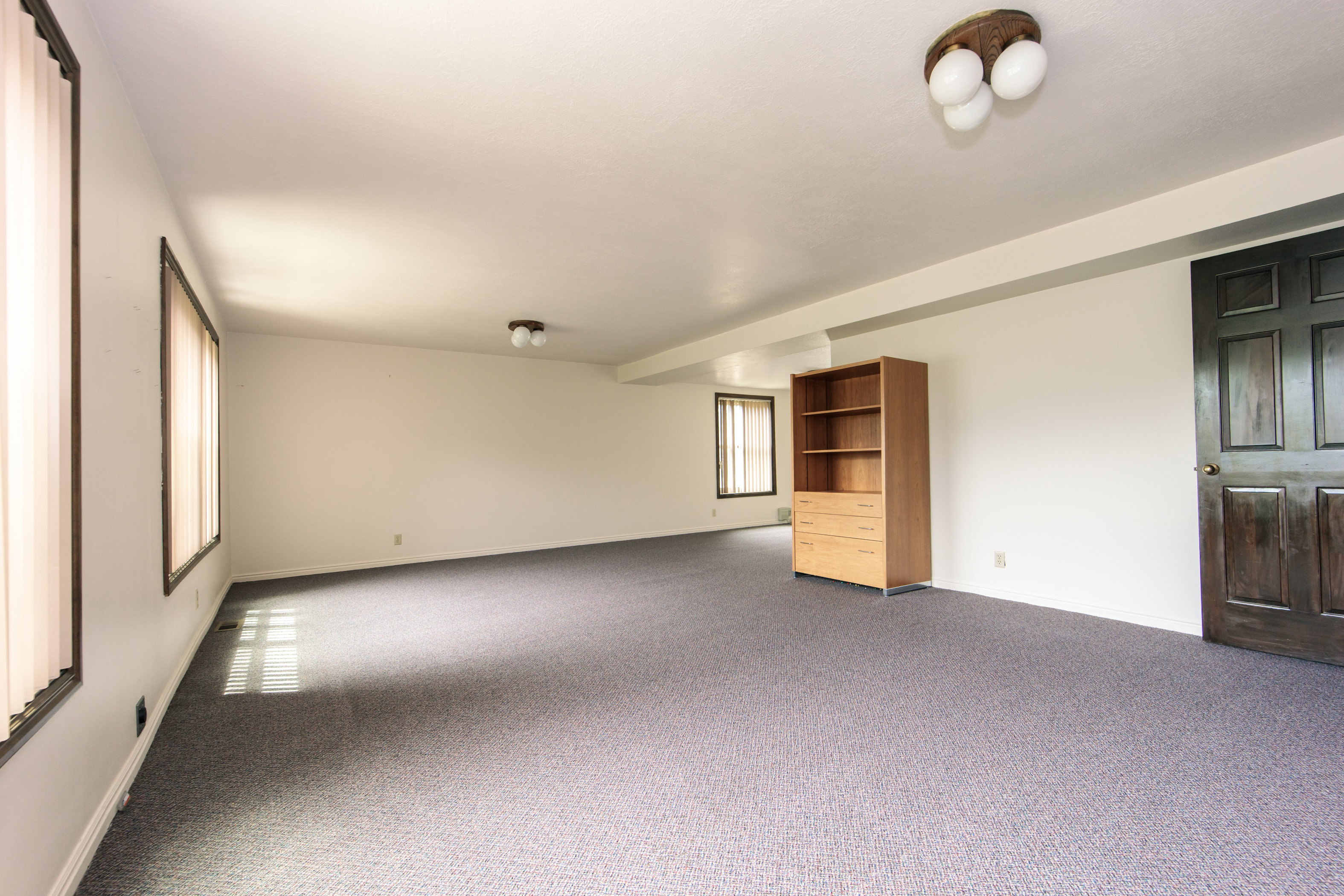 Spacious room featuring gray textured carpeting, white walls, and multiple windows with vertical blinds