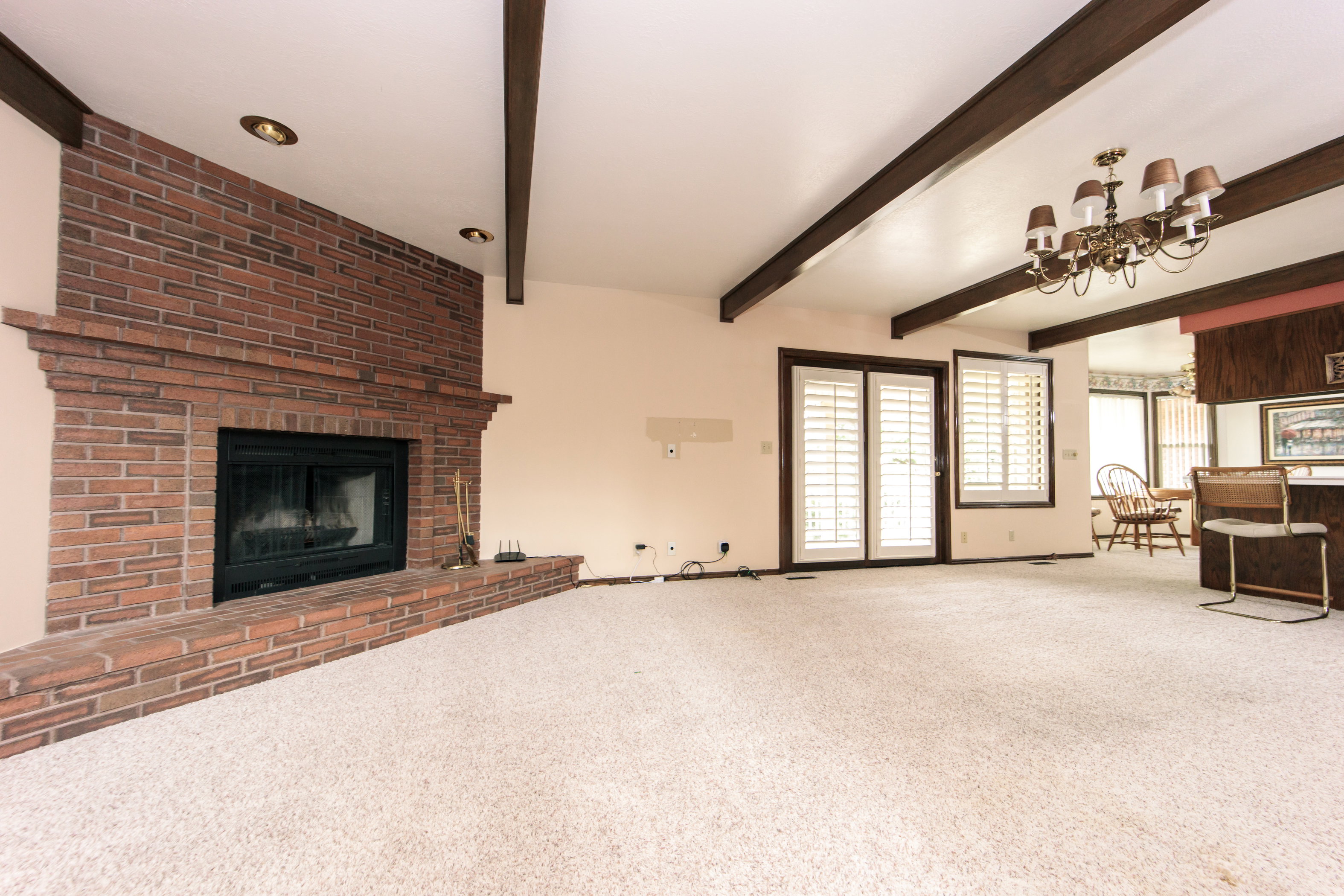 Living area featuring a brick fireplace with a raised hearth, exposed wood ceiling beams, recessed lighting, and a brass chandelier