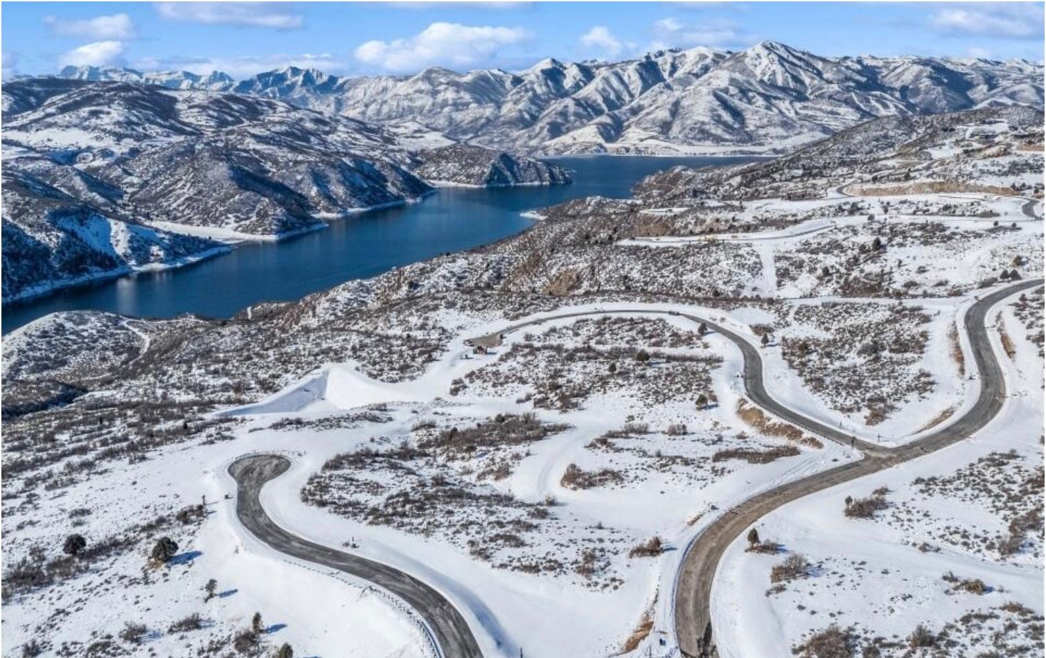 Expansive snow-covered landscape featuring multiple winding roads and a prominent body of water bordered by snow-capped mountains