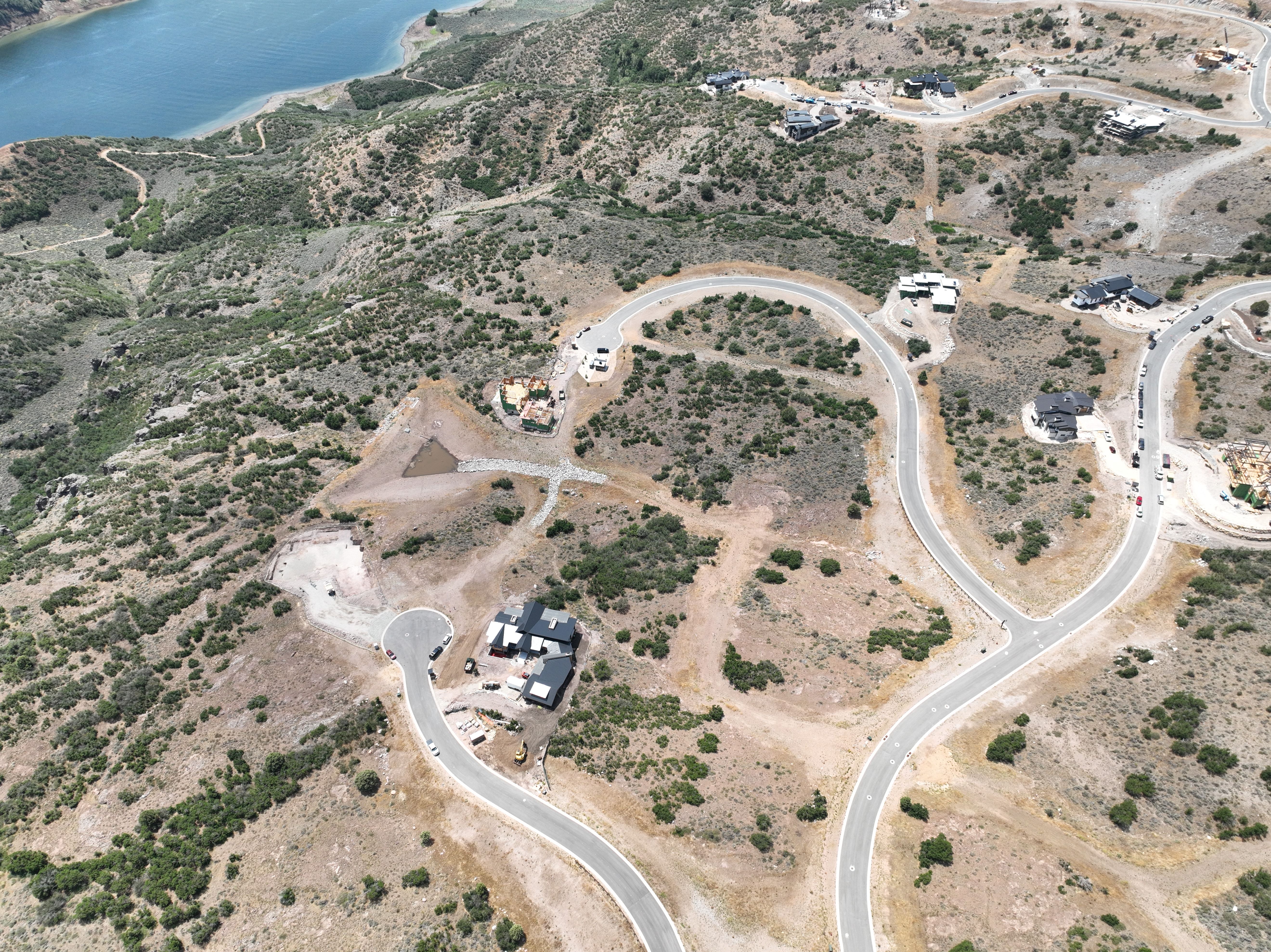 Aerial perspective of a hillside residential development featuring a lake, winding paved roads, established homes, and active construction sites