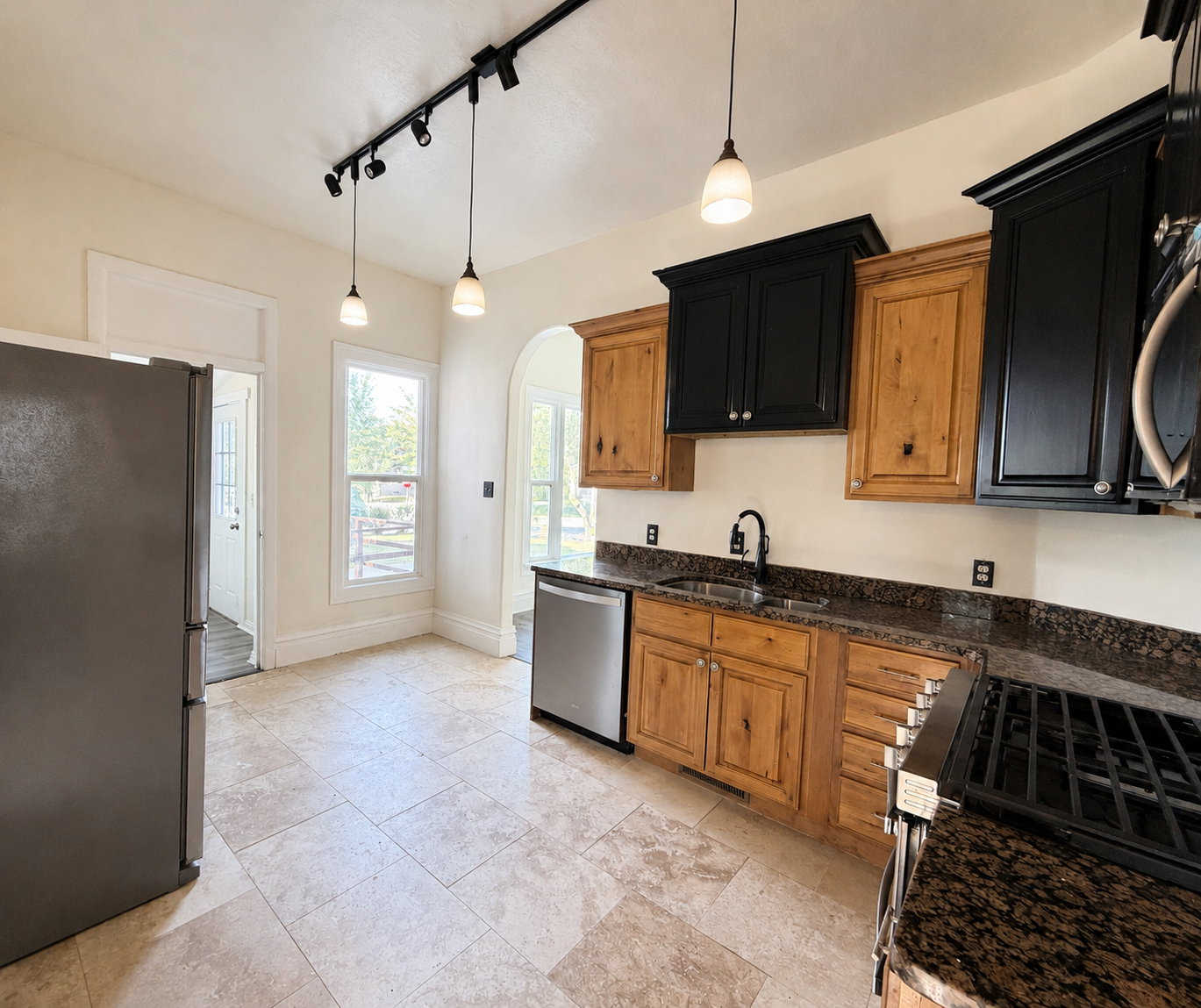 Kitchen featuring tile flooring, dark stone countertops, wood cabinetry with black upper cabinets, a stainless steel dishwasher, and a stainless steel refrigerator