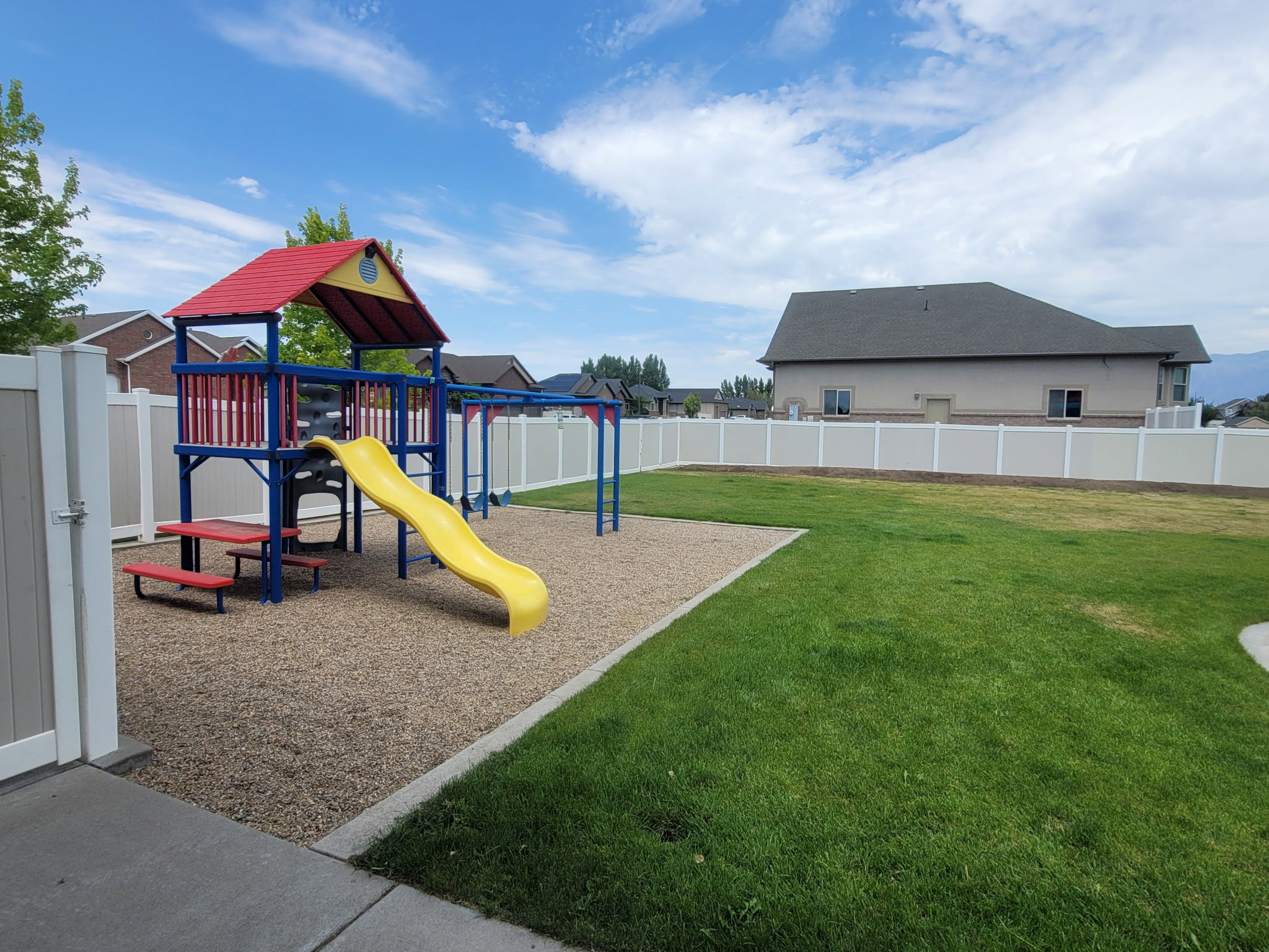 Expansive backyard featuring a colorful playground structure with slides and swings, wood chip ground cover, a large grassy area, and a privacy fence