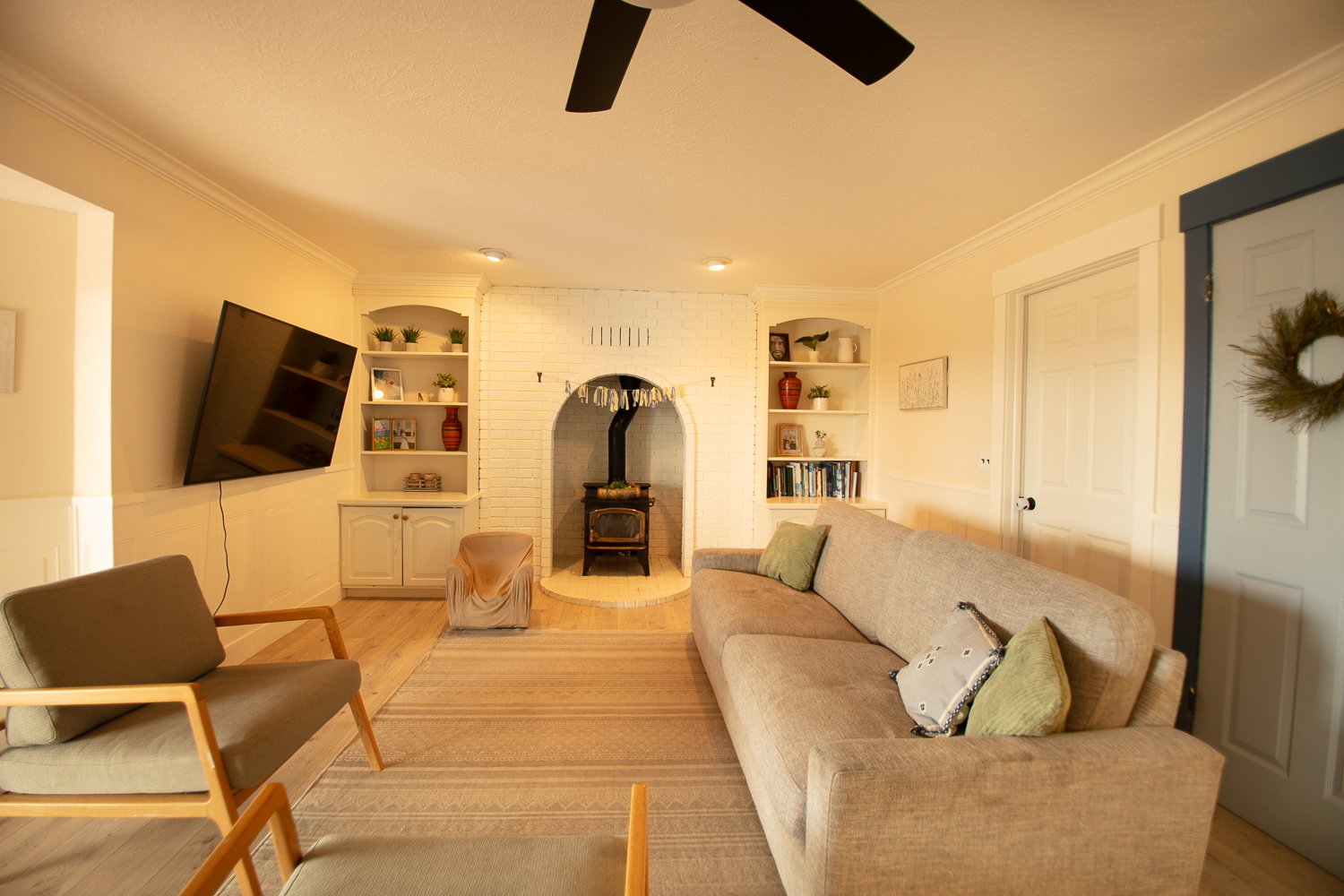 Living area featuring a white-painted brick fireplace with a wood-burning stove, built-in shelving, wood-finish flooring, and crown molding