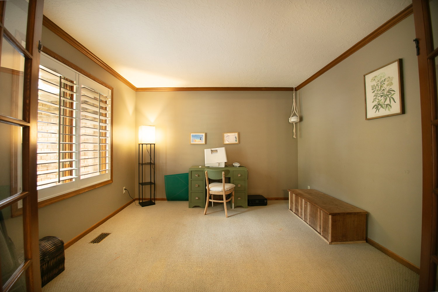 Room featuring a window with plantation shutters, wood-finish trim, neutral wall paint, and light-colored carpet flooring