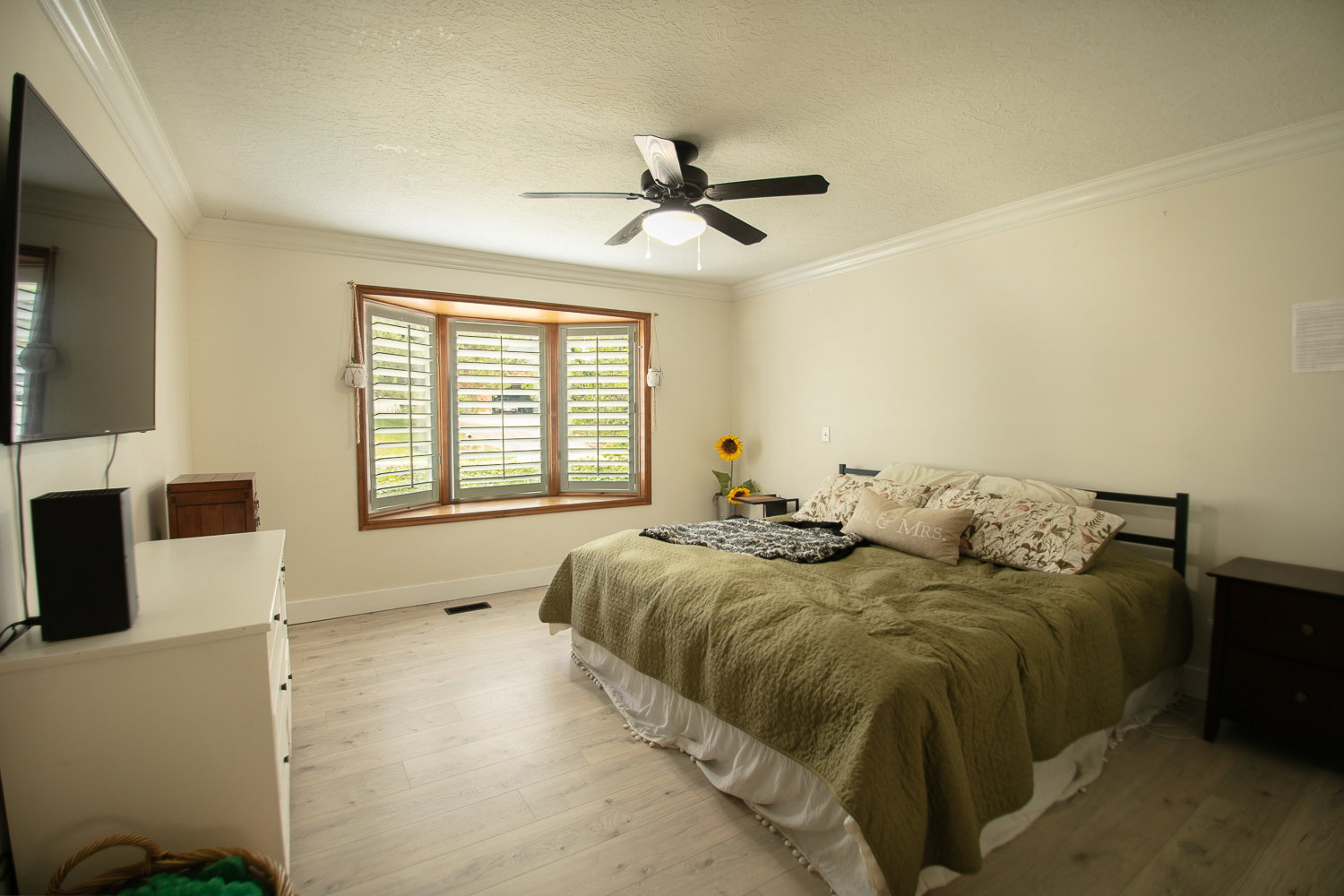 Spacious room featuring a multi-pane bay window with louvered shutters, crown molding, a ceiling fan, and light wood-finish flooring