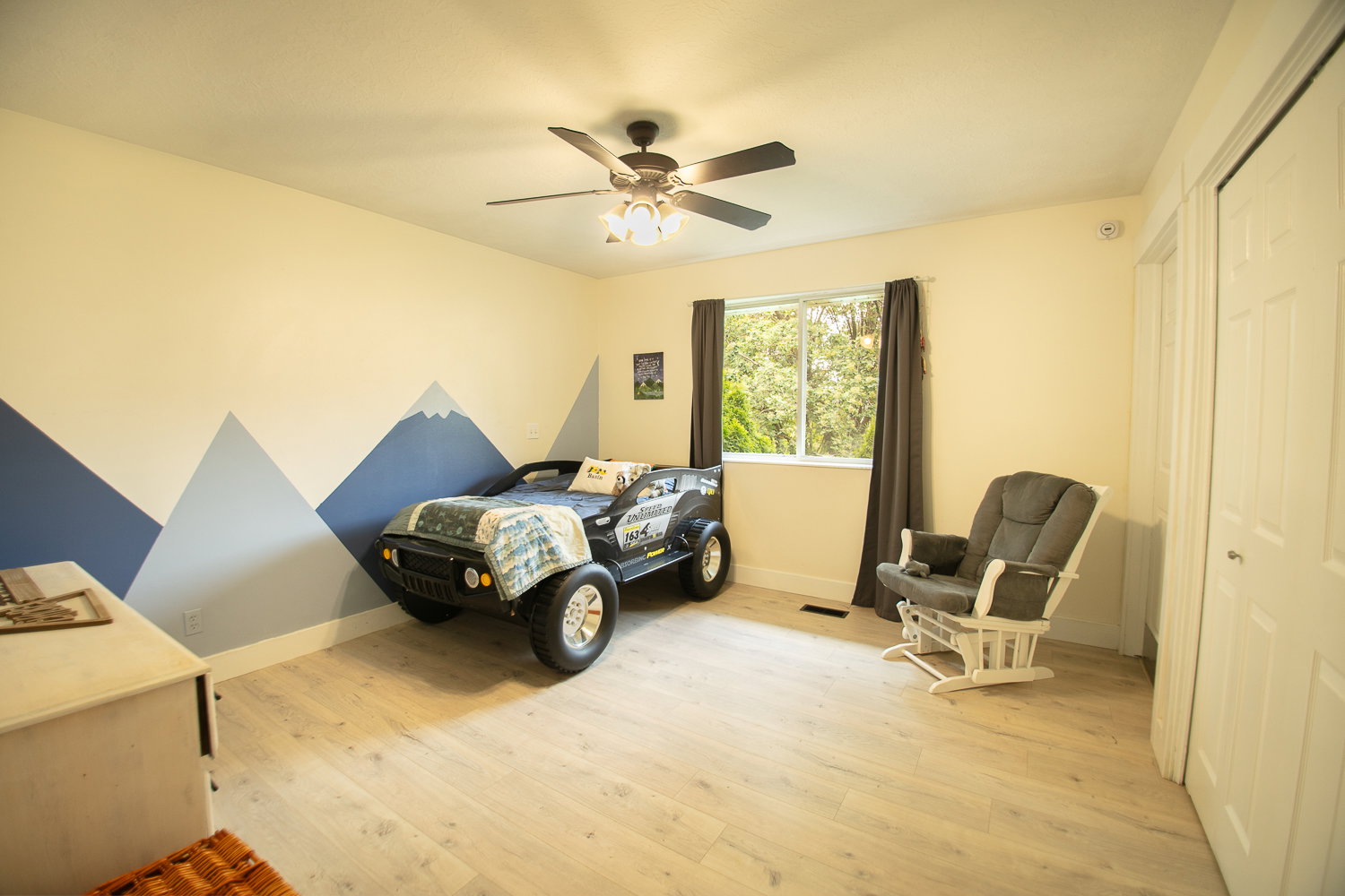 Bedroom featuring light wood-finish flooring, a ceiling fan with integrated lighting, and a window with dark curtains