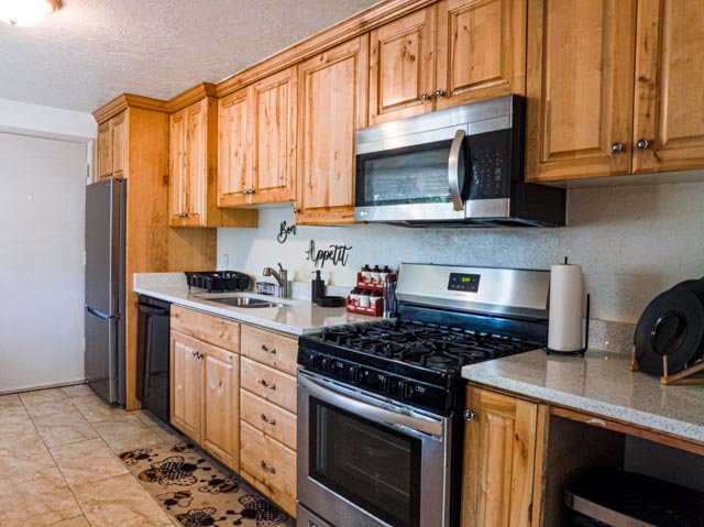 Kitchen featuring natural wood cabinetry, stainless steel appliances, light-toned countertops, tile flooring, and a white tile backsplash