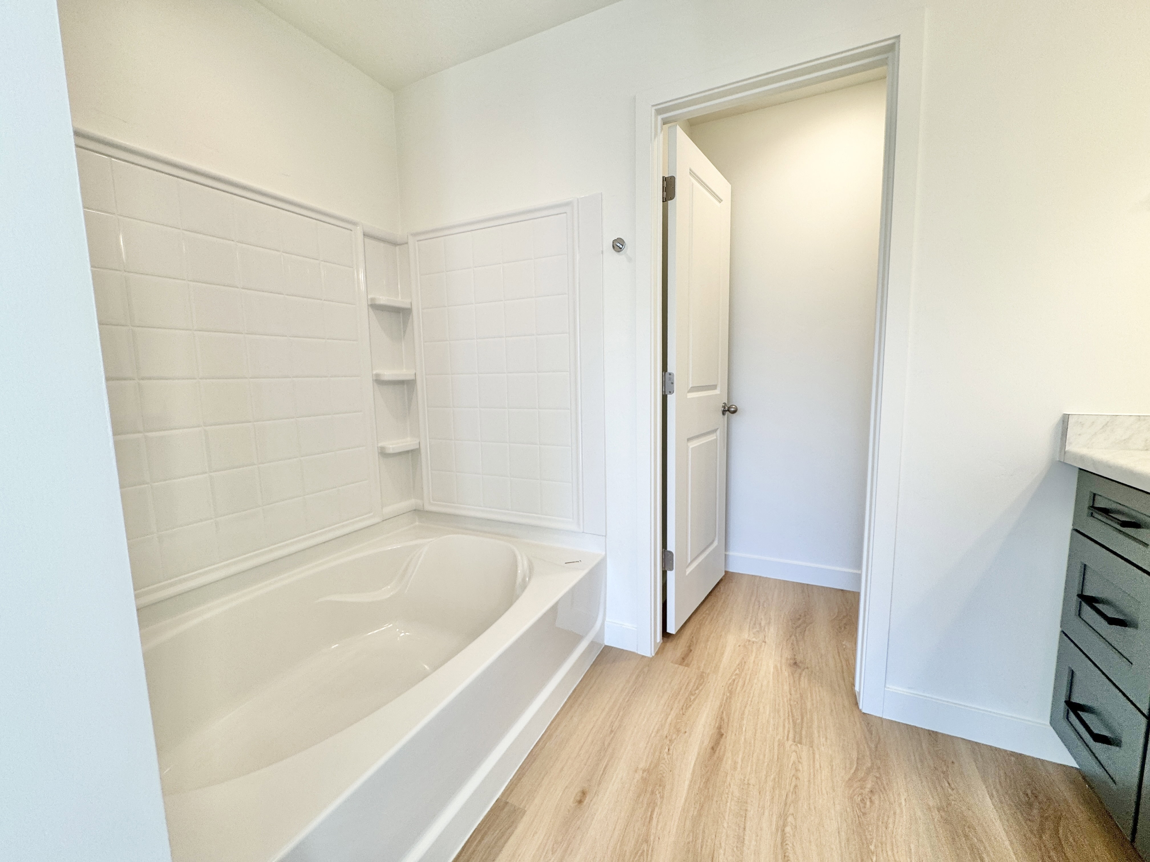 Bathroom featuring a built-in tub with white tile surround and integrated shelving, wood-finish flooring, and a vanity with a light-colored countertop and dark green cabinetry