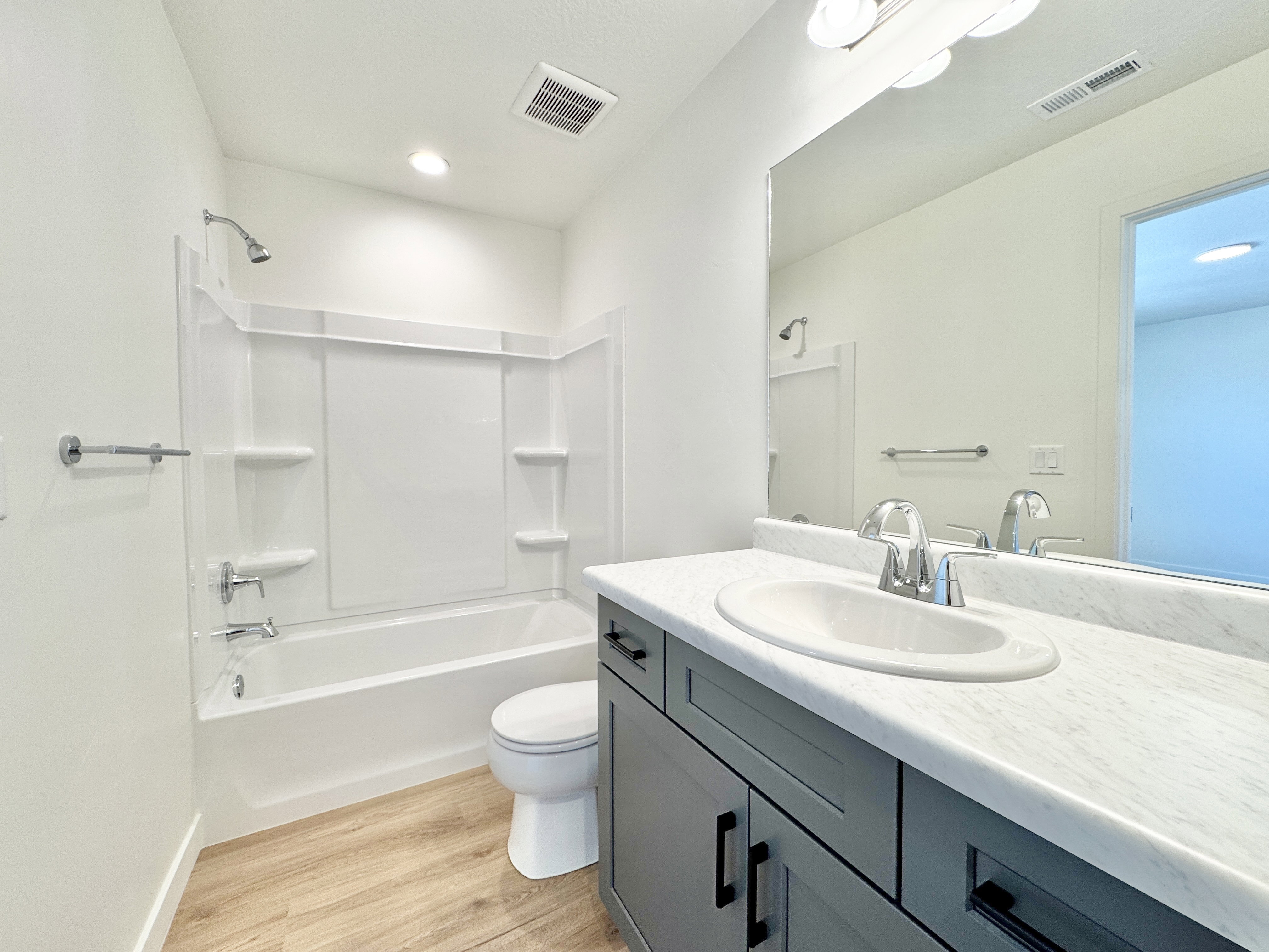 Bathroom featuring a white tub/shower combination with chrome fixtures, a dark gray vanity with black hardware, a white undermount sink, a white countertop, and light wood-finish flooring