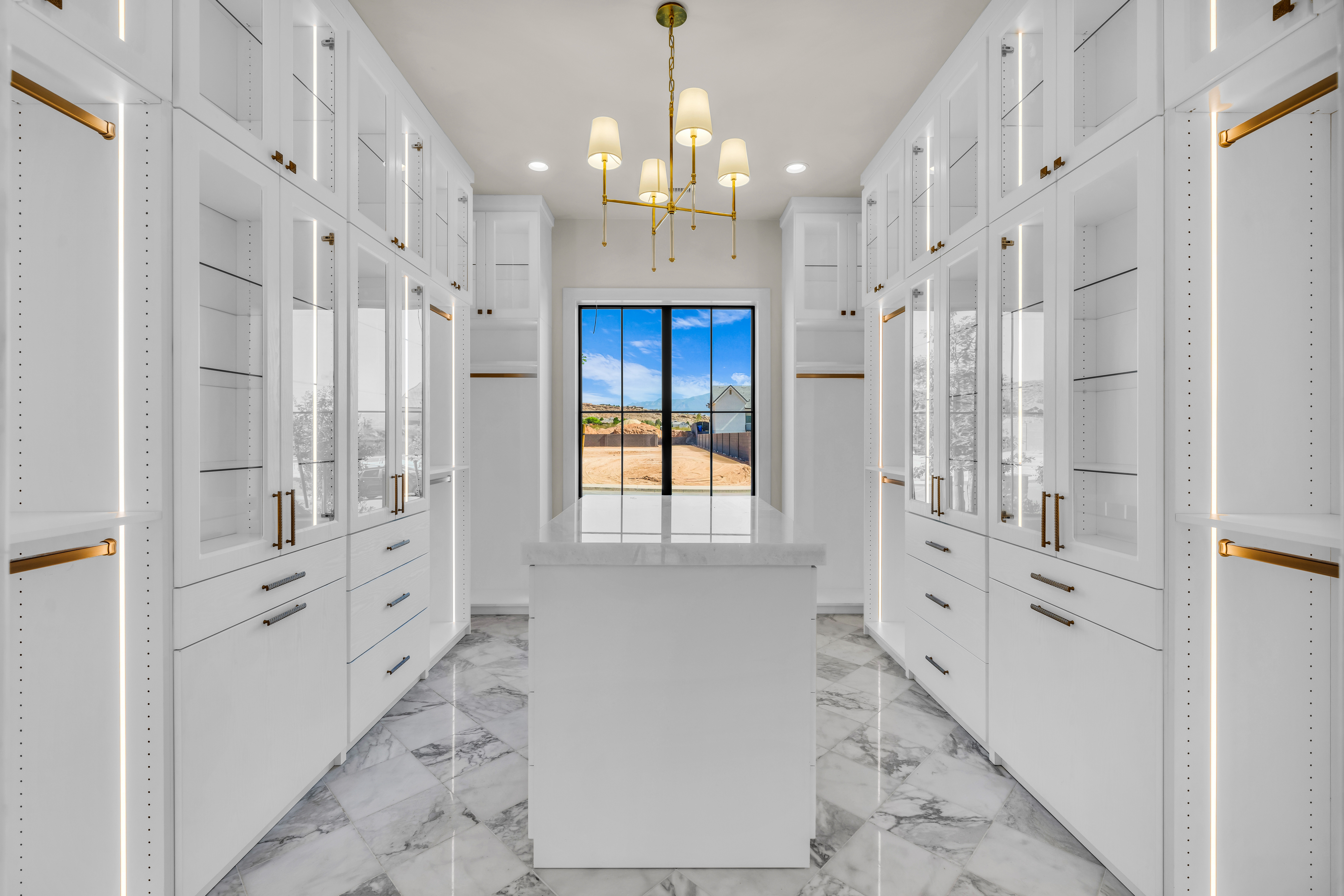 Spacious custom closet featuring extensive white cabinetry with glass-front uppers, an integrated center island, brass hardware, and interior lighting
