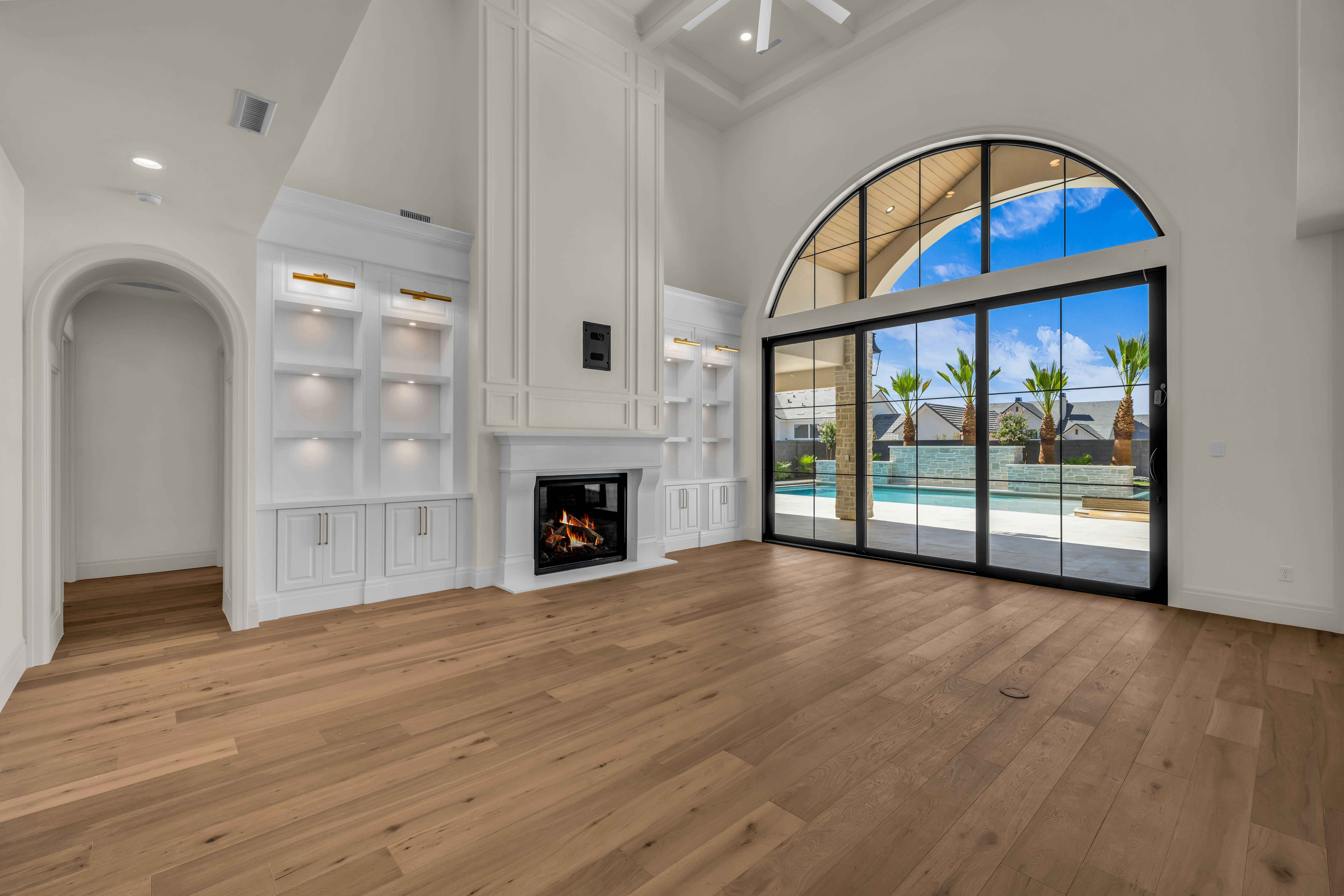 Great room featuring high ceilings, wood-finish flooring, a built-in fireplace with a mantel, integrated shelving with accent lighting, and an arched window wall with sliding glass doors