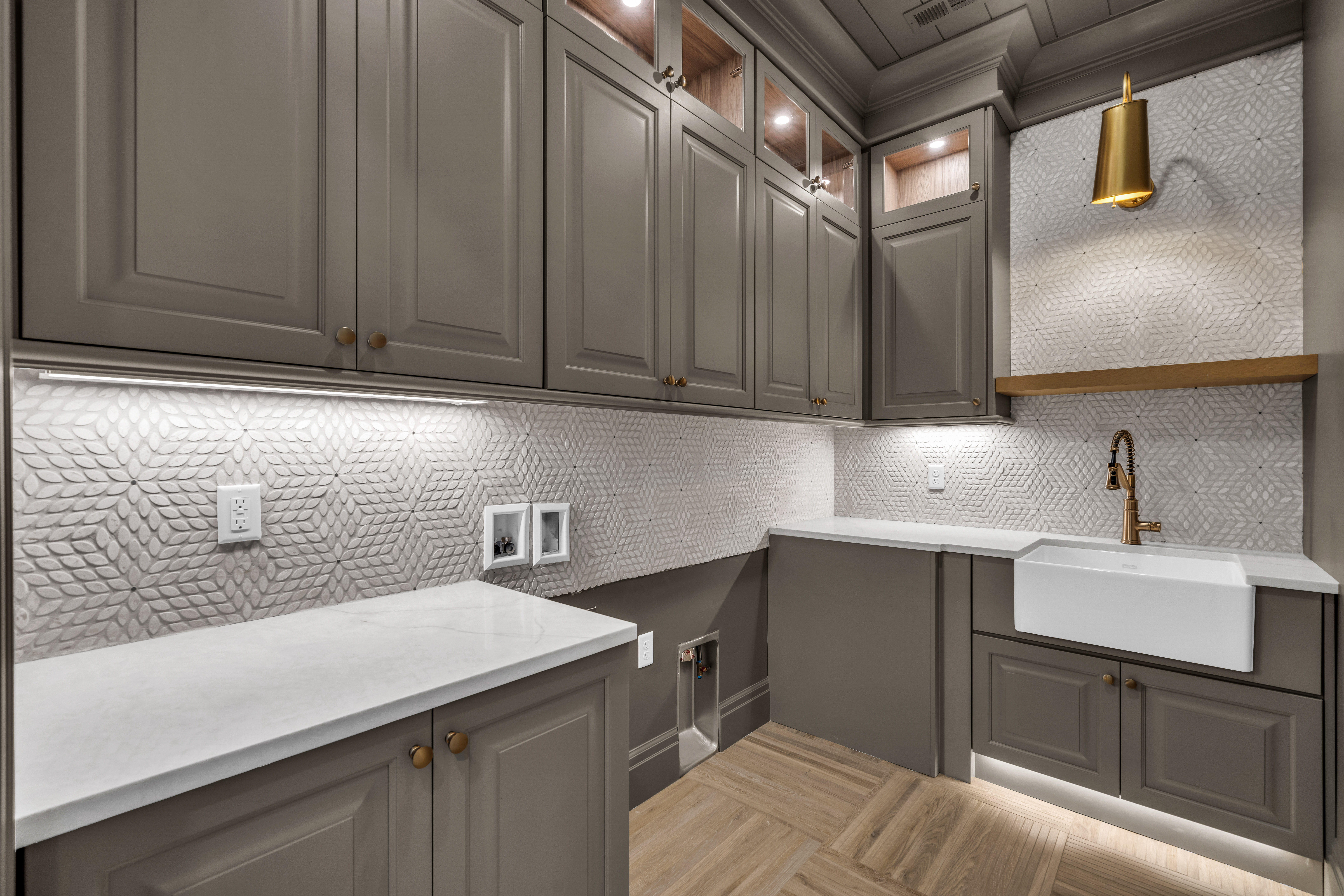 Laundry room featuring light gray cabinetry with gold-toned hardware, white quartz countertops, and a white farmhouse sink with a brass faucet