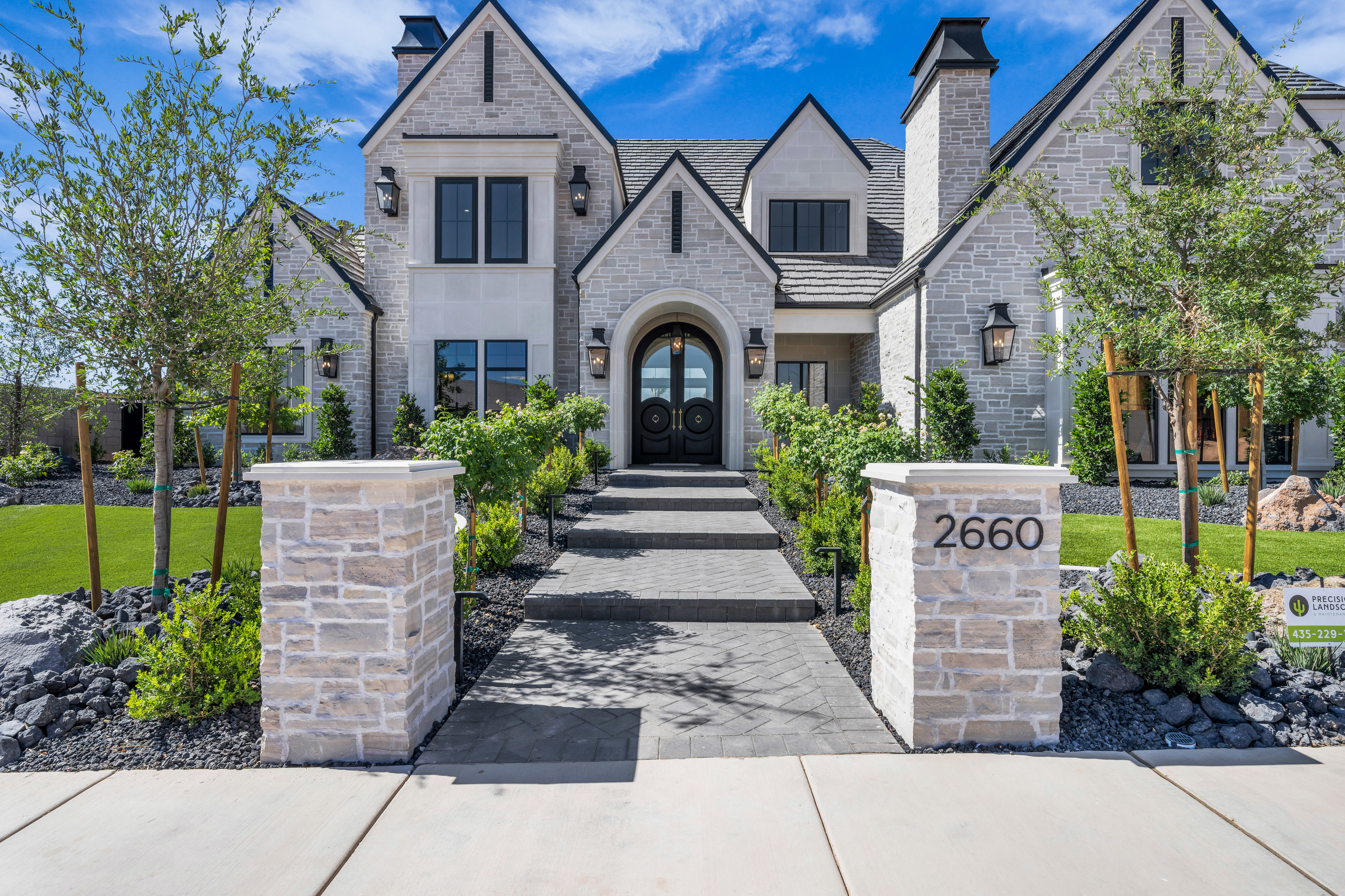 Stone-clad exterior featuring multiple gables, dark-framed windows, a prominent arched entry, brick chimneys, and an inviting walkway