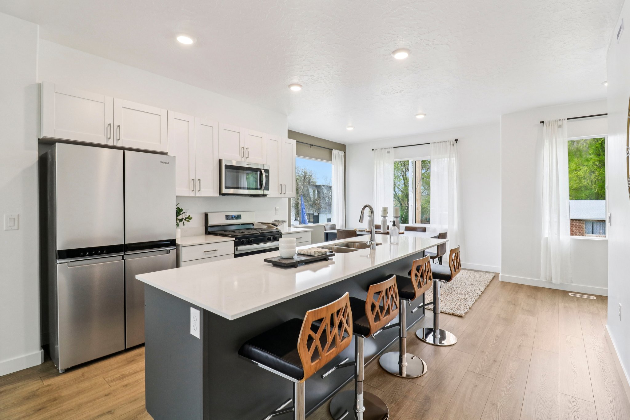 Open-concept kitchen featuring an oversized island with a dual-basin sink, stainless steel appliances, white cabinetry, recessed lighting, and wood-finish flooring