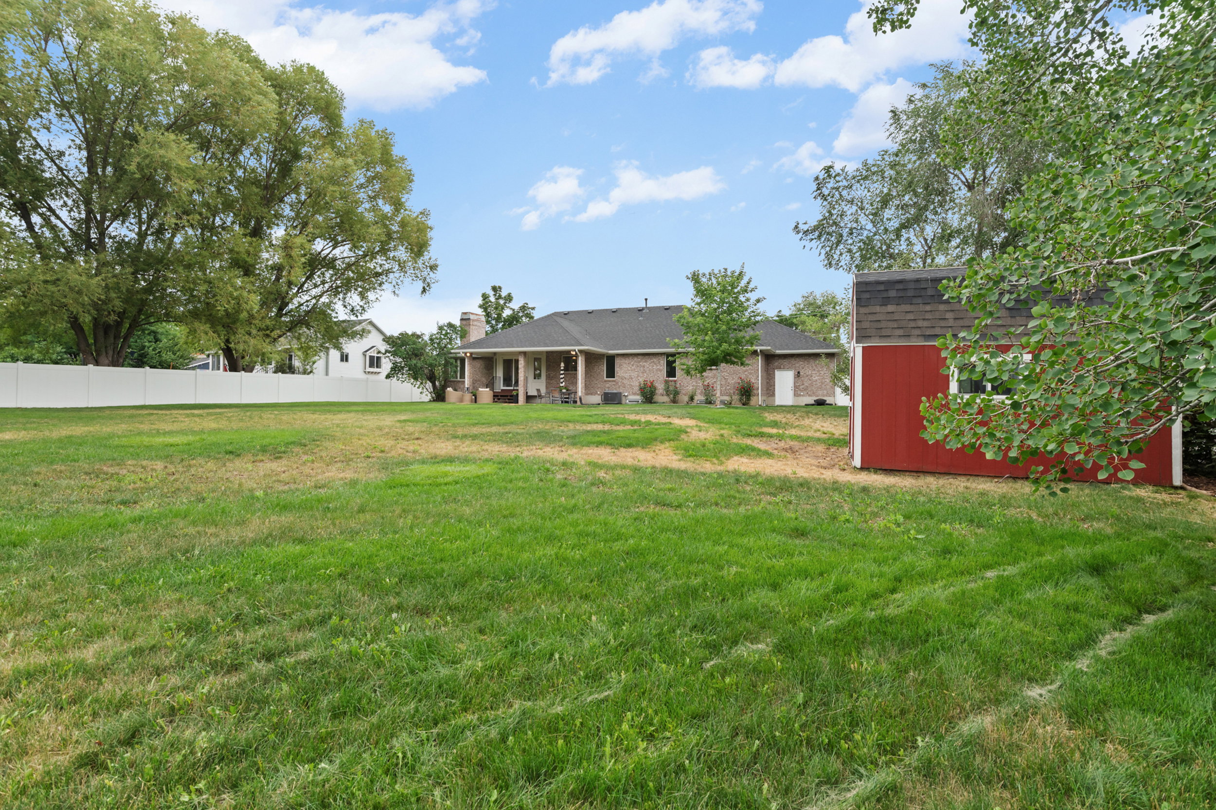 Spacious backyard featuring a manicured lawn, a detached red storage shed with a dark shingle roof, mature perimeter trees, and a white privacy fence