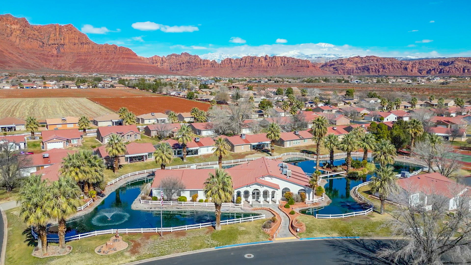Community aerial featuring a central building surrounded by decorative waterways with fountains, mature palm trees, and perimeter fencing