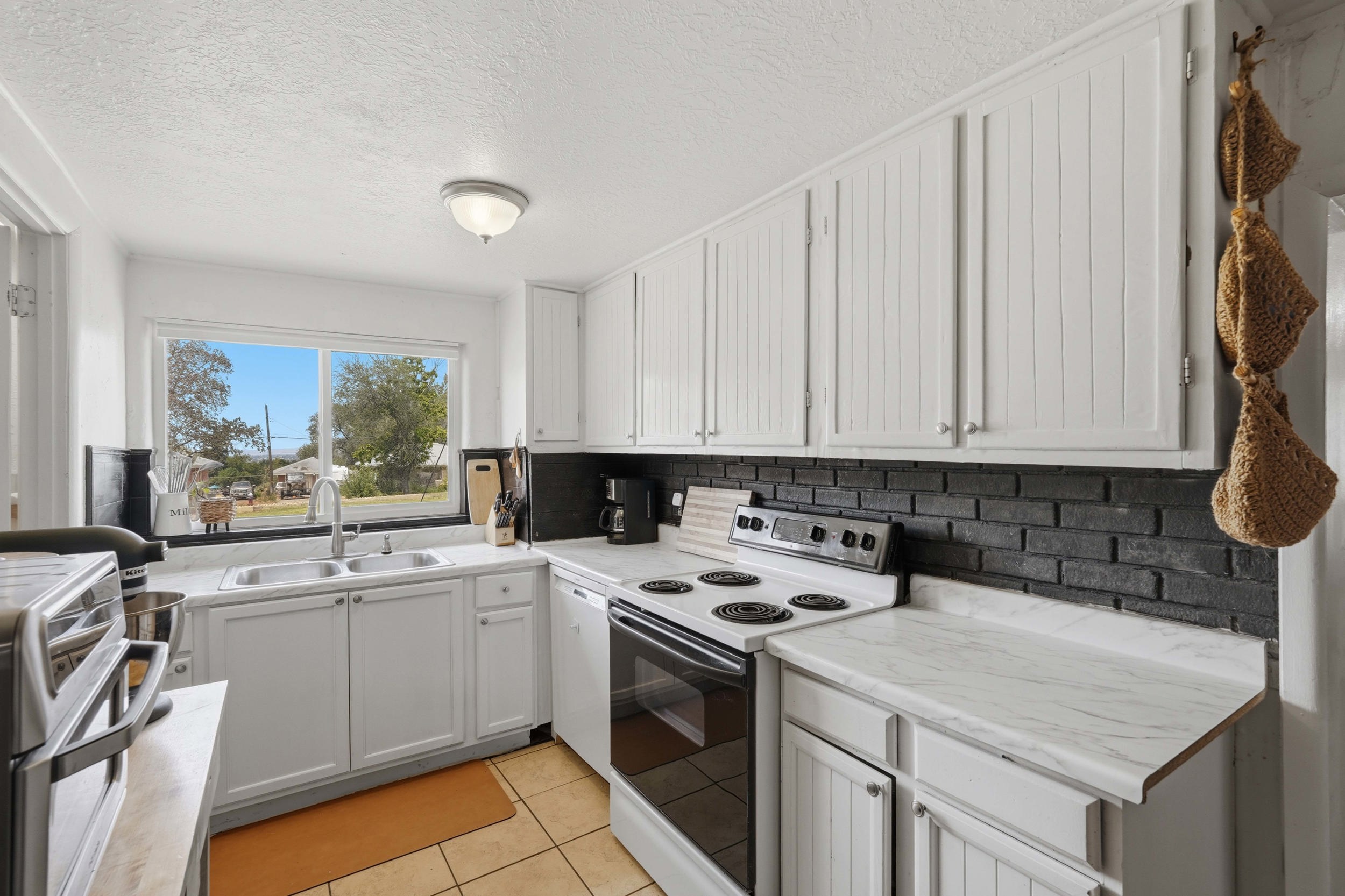 Bright kitchen featuring a double basin stainless steel sink, white cabinetry, a black brick-style backsplash, and light-toned countertops