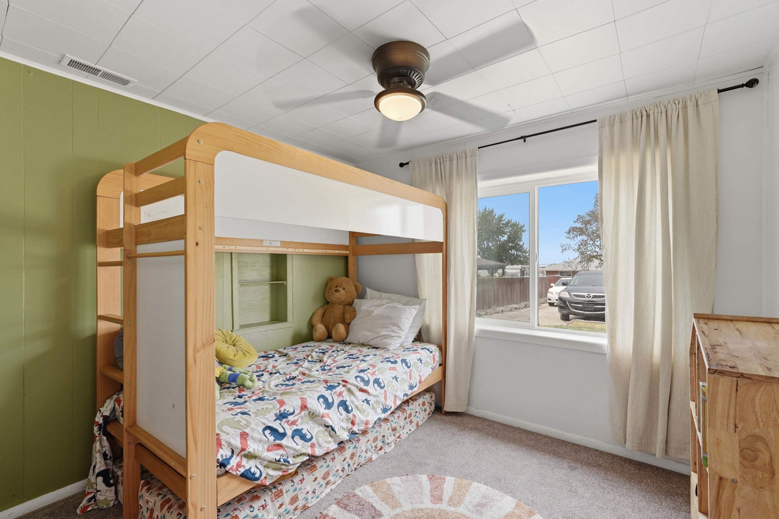 Room featuring a window with natural light, green paneled accent wall, neutral carpet flooring, and a ceiling fan with integrated lighting