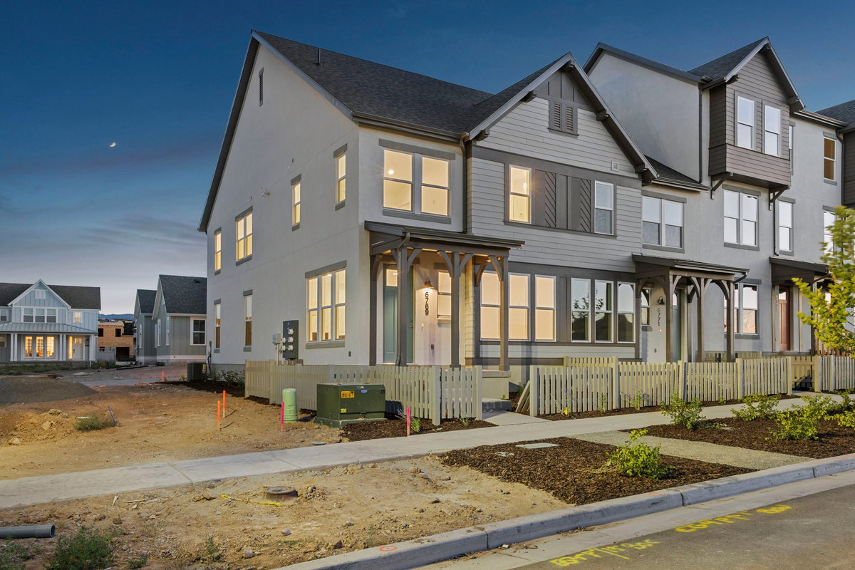 Modern townhome exterior featuring a light gray and white facade, dark gray gabled roof, multiple windows with white trim, and a decorative picket fence