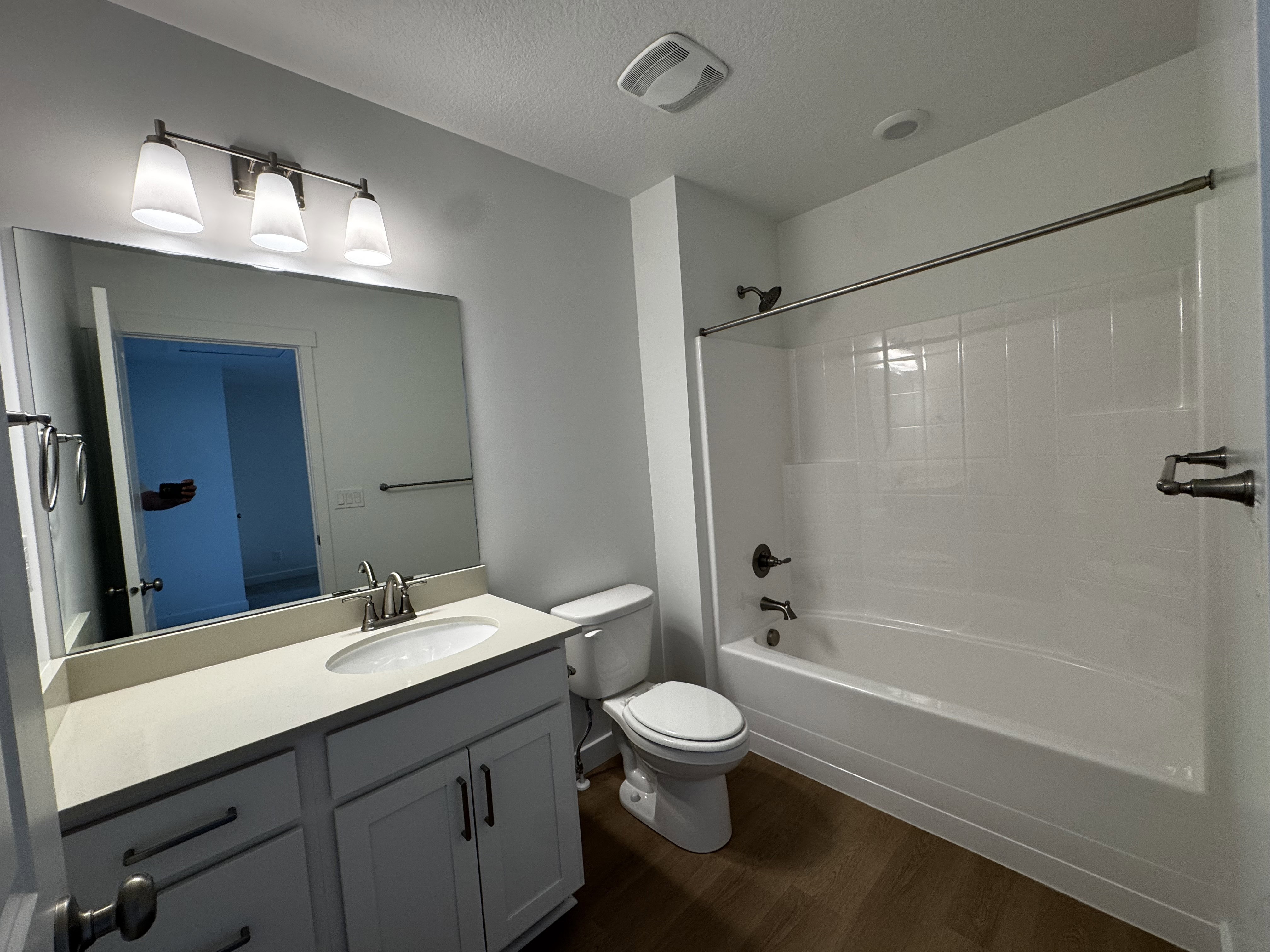 Contemporary bathroom featuring a single vanity with a light-toned countertop, a built-in tub/shower combination, dark bronze fixtures, a wall-mounted light fixture, and wood-finish flooring