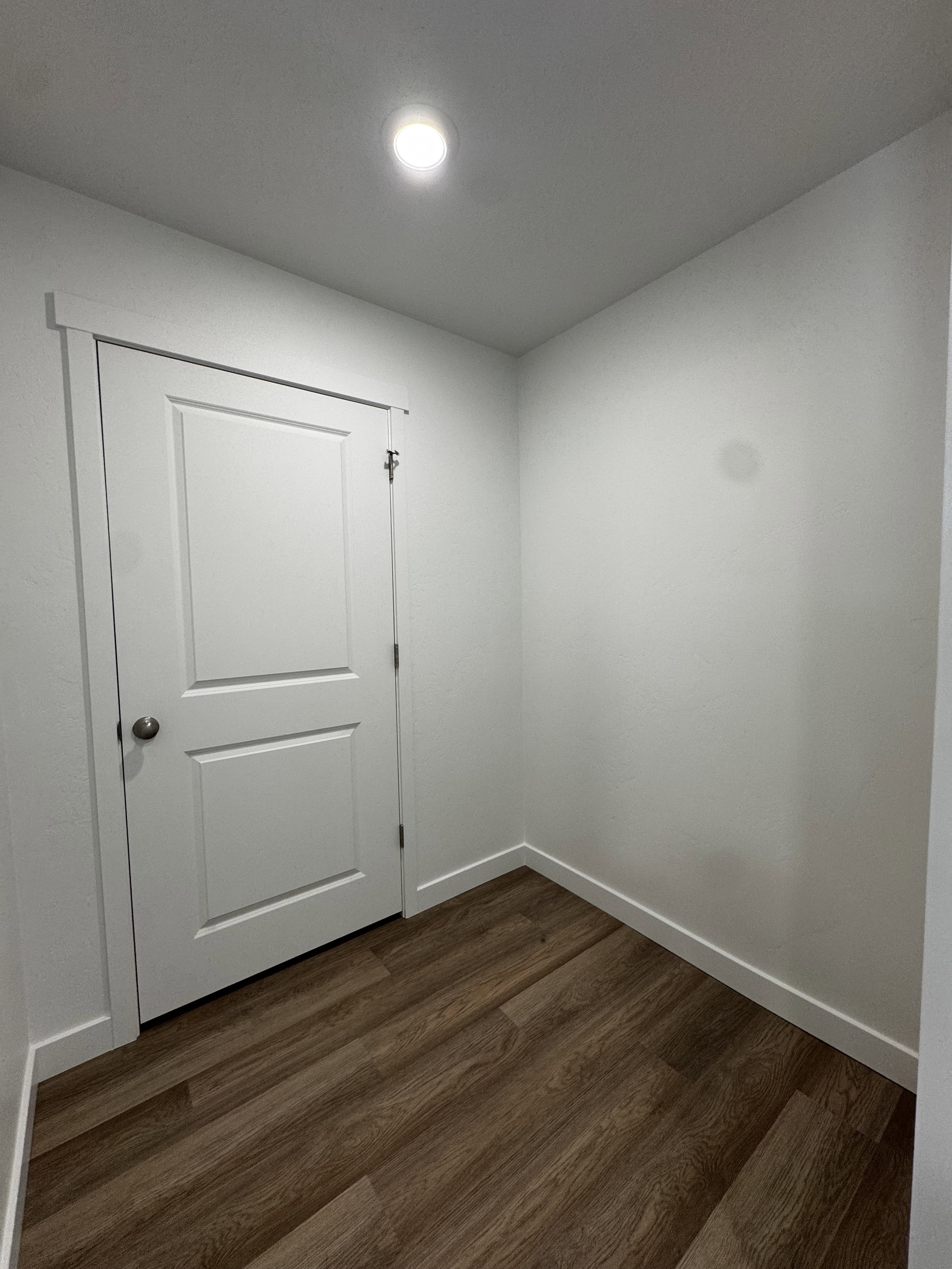 Recessed lighting illuminates wood-finish flooring, white walls, and a white paneled door with a round silver-tone knob