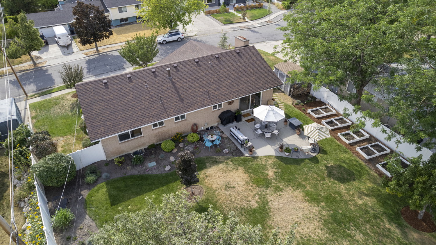 Expansive backyard featuring a curved concrete patio, established raised garden beds, a mature tree canopy, and a durable shingled roof