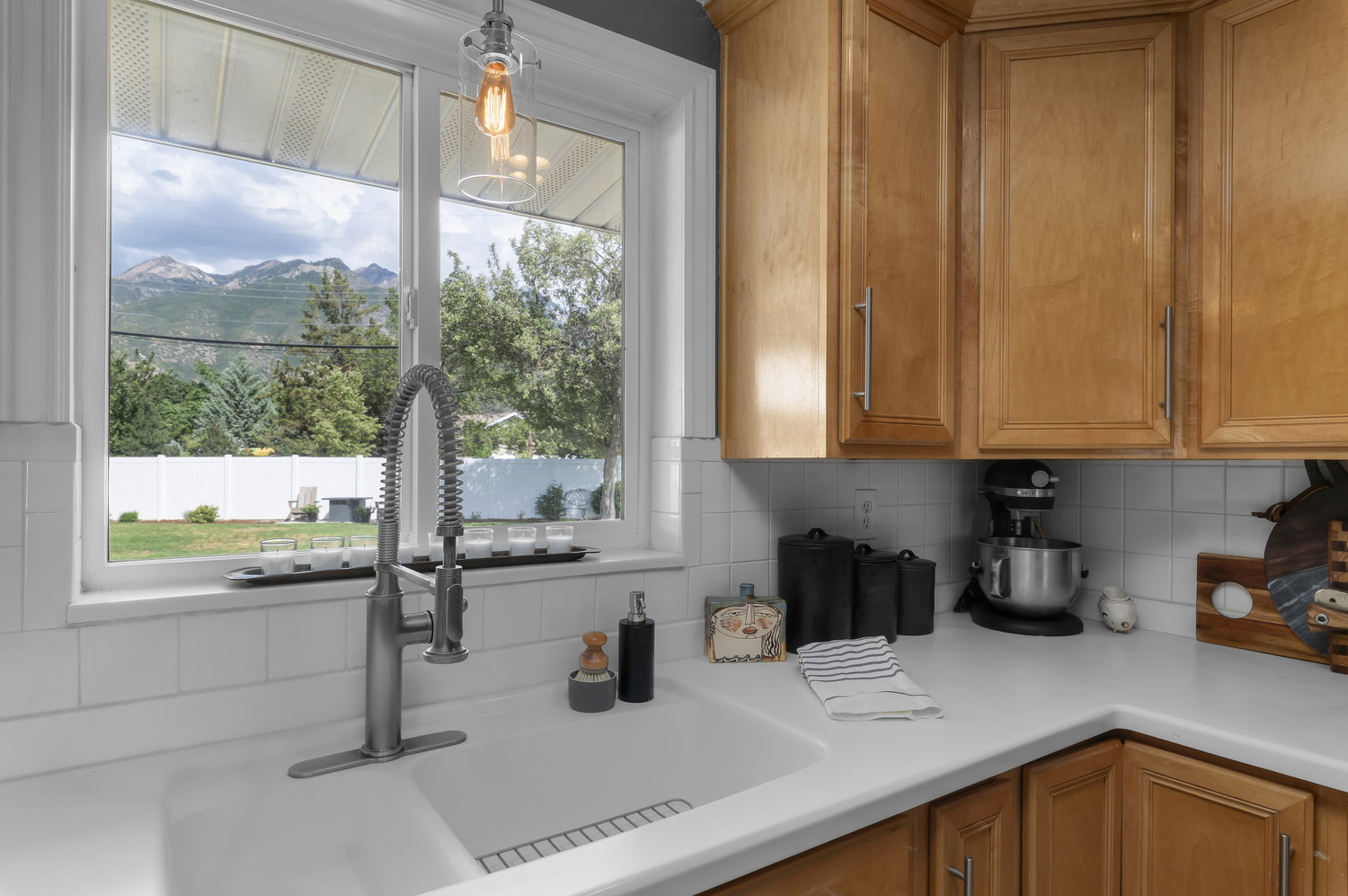 Kitchen sink with a contemporary pull-down faucet, white solid surface countertop, light wood cabinetry, and a subway tile backsplash