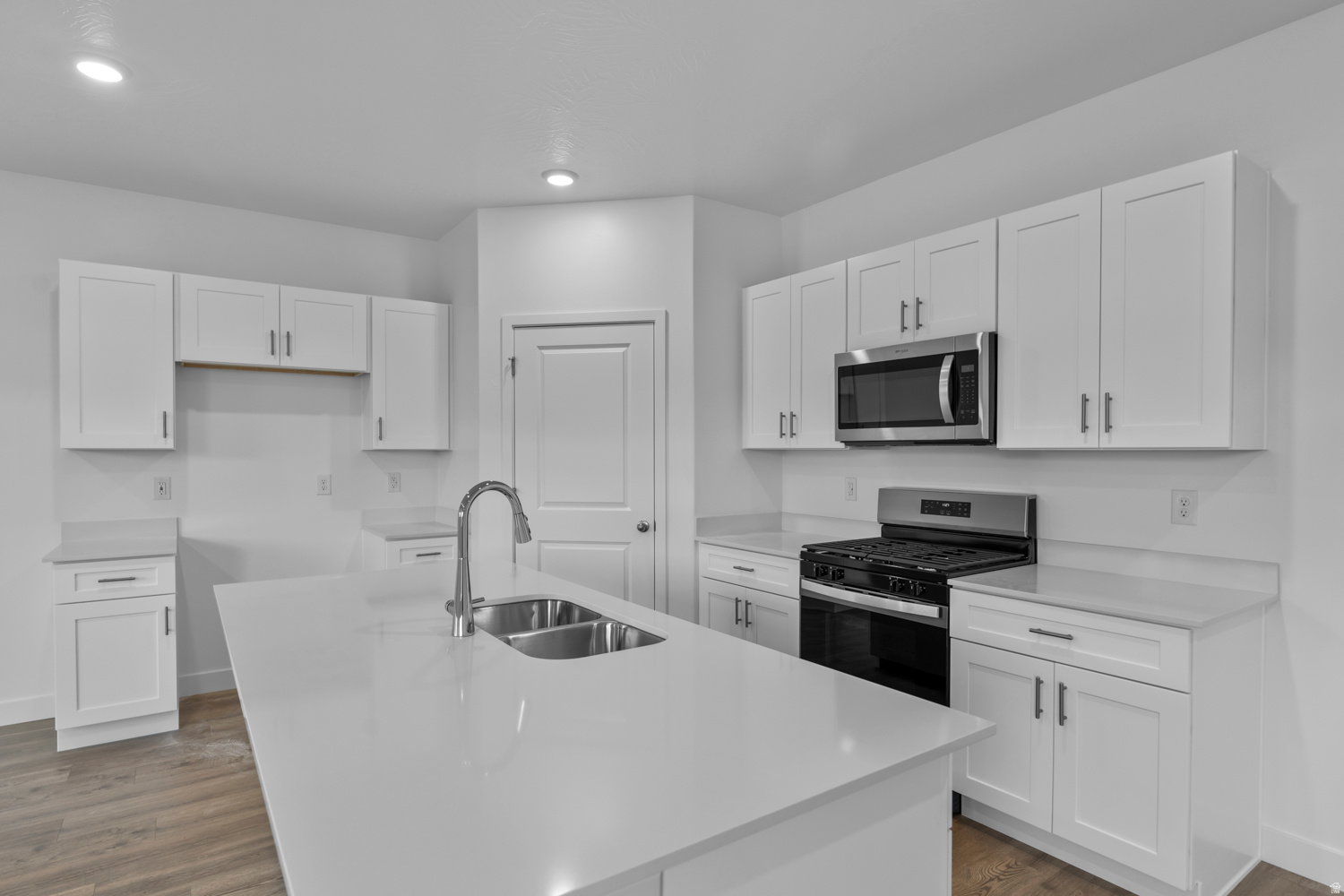 Kitchen featuring white shaker-style cabinetry, a central island with a double basin sink and gooseneck faucet, light-toned countertops, stainless steel appliances, and wood-finish flooring