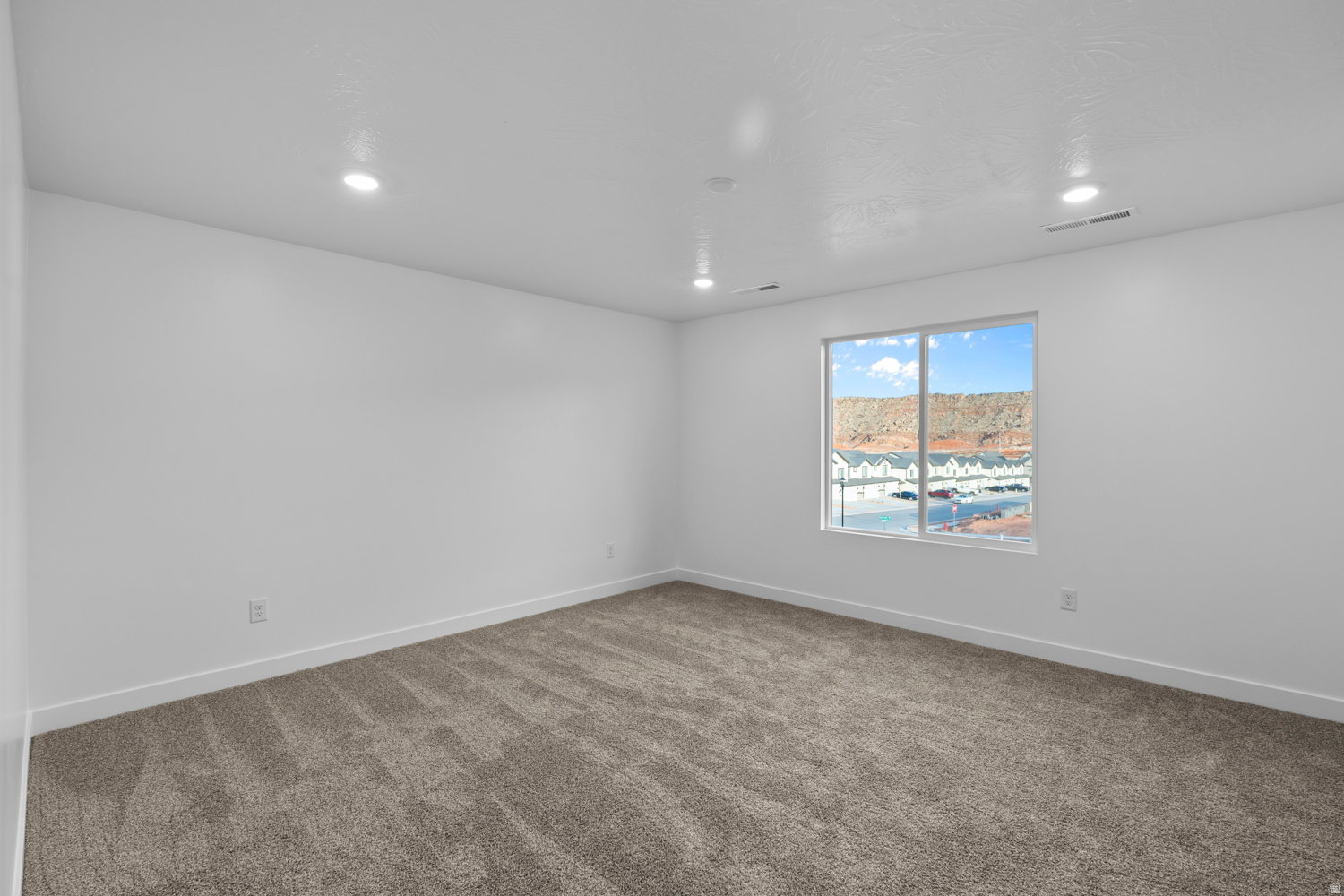 Spacious room featuring neutral carpeting, white walls, recessed ceiling lighting, and a large window providing natural illumination and a distant mountain backdrop