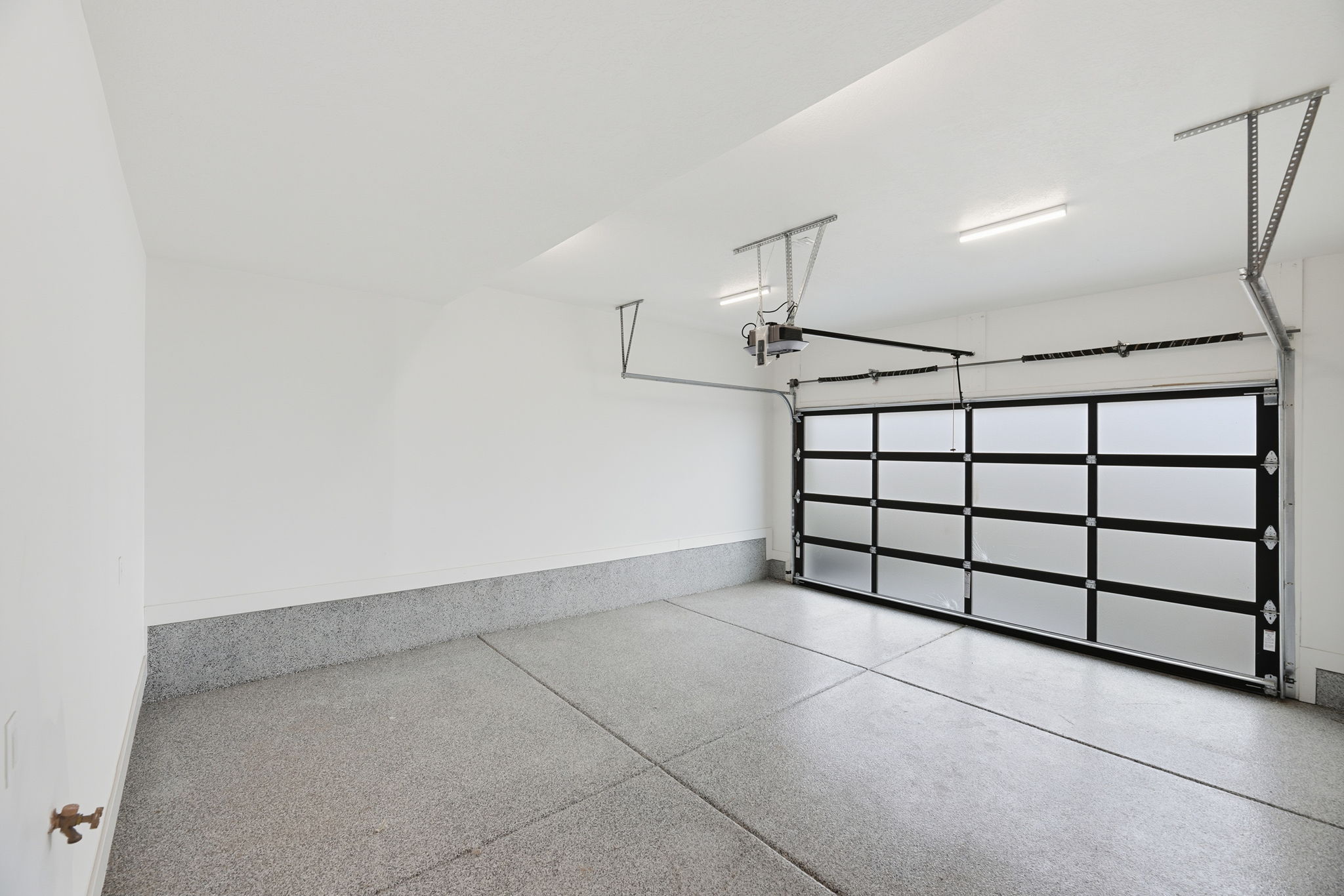 Spacious two-car garage featuring a modern frosted glass and black frame garage door, epoxy-coated concrete flooring, bright overhead lighting, high ceilings, and an integrated wall spigot