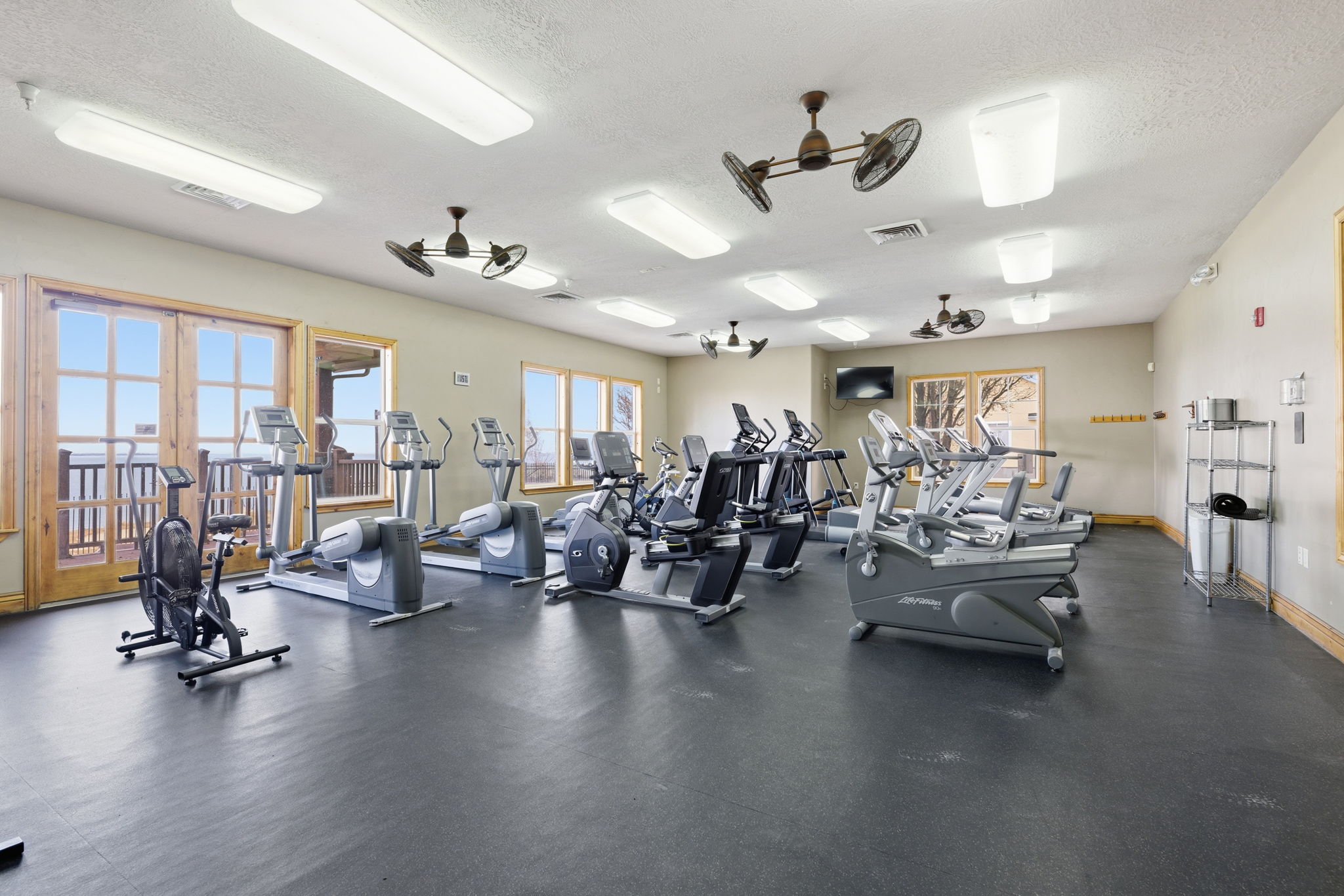 Spacious fitness room featuring rubber flooring, multiple cardio machines, ceiling-mounted fans, recessed lighting, and wood-framed windows