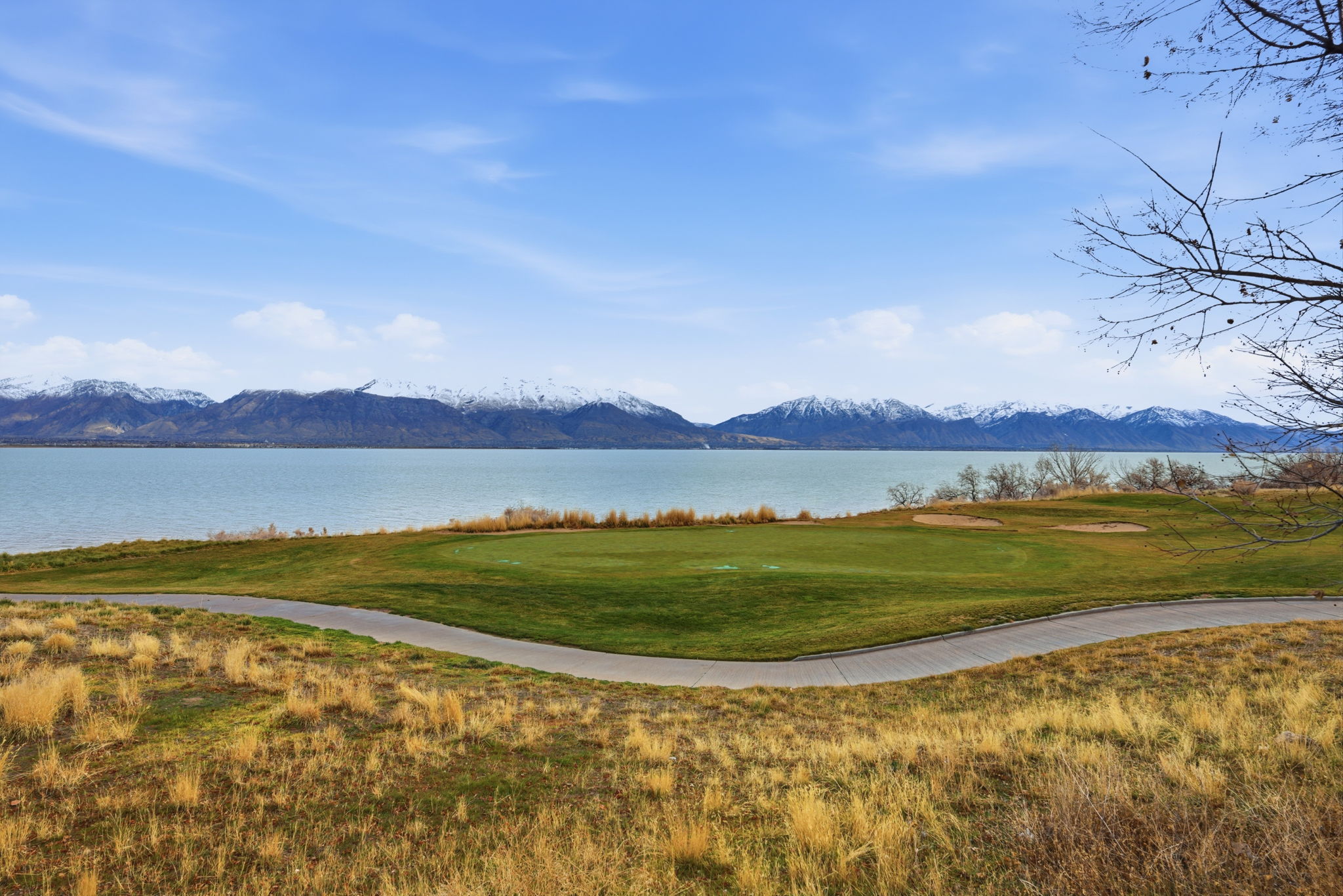 Expansive golf course green with sand traps bordering a large body of water, set against a backdrop of snow-capped mountains under a clear blue sky