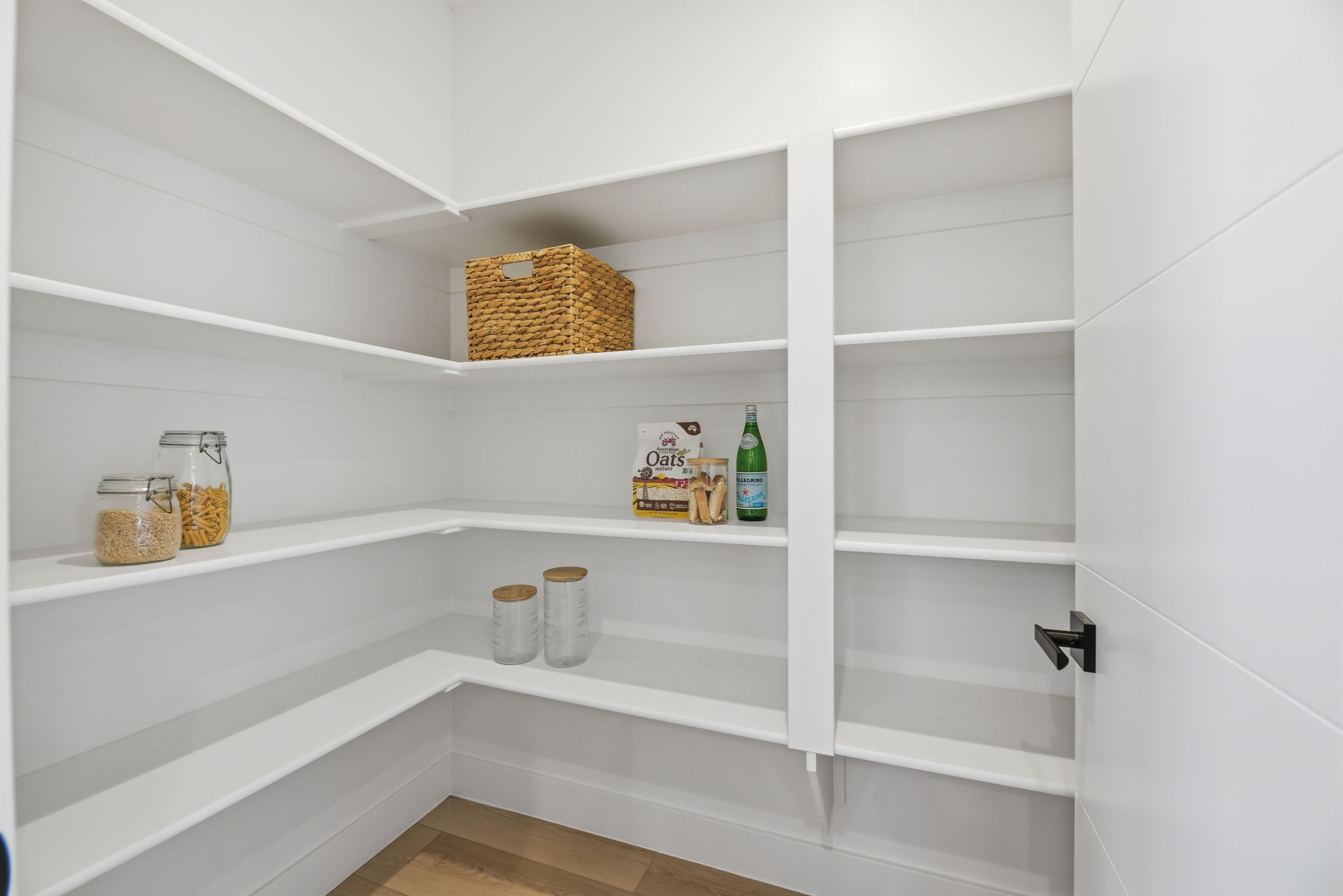 Spacious walk-in pantry featuring extensive white shelving, wood-finish flooring, and a contemporary matte black door handle