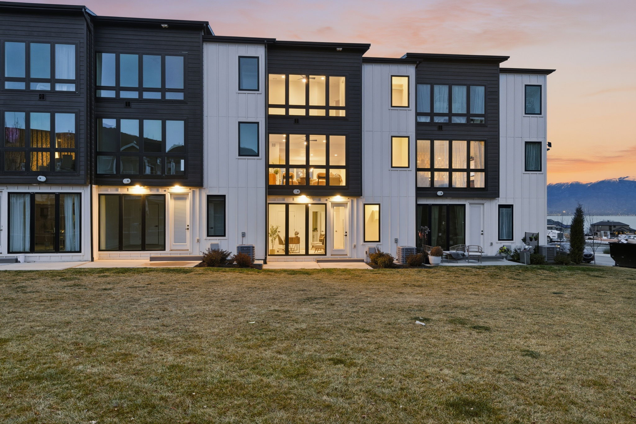 Modern townhome exteriors featuring contrasting dark and light siding, multiple large windows, ground-floor patios, and a grassy backyard area