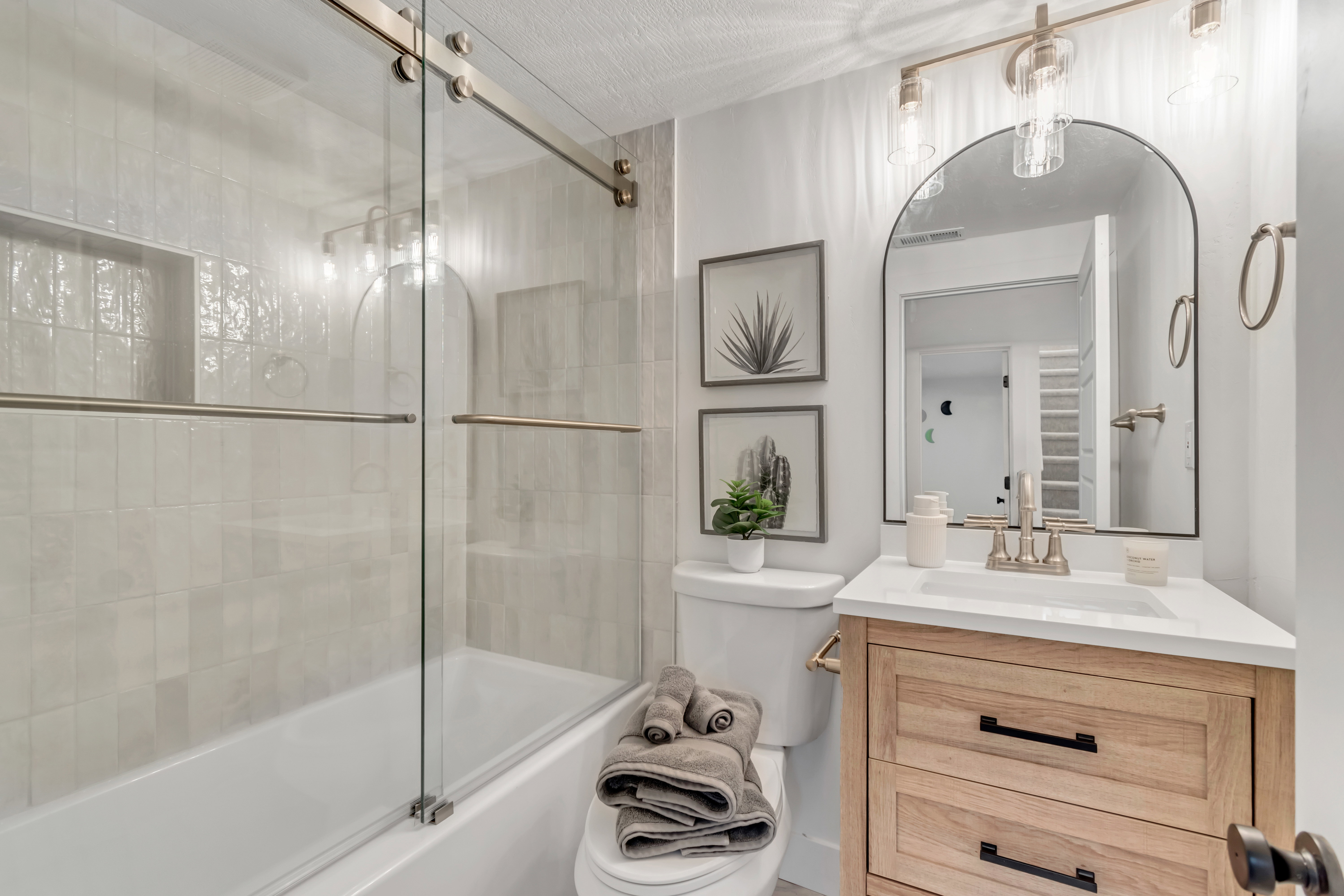 Bathroom featuring a wood-finish vanity with a white countertop and rectangular undermount sink, a large arched mirror, and a full-size bathtub with a glass sliding enclosure