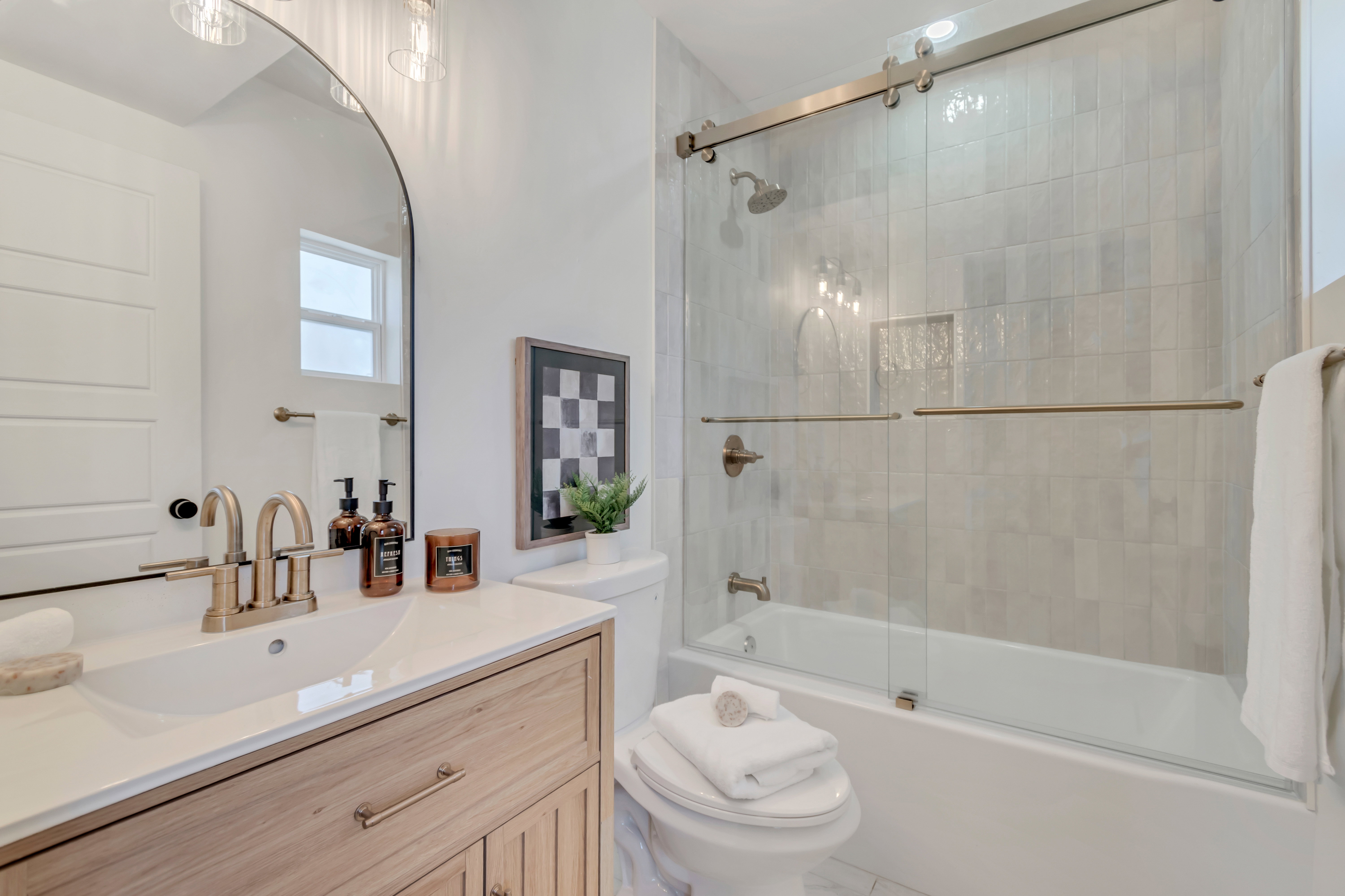 Bathroom featuring a wood-finish vanity with a white countertop and rectangular undermount sink, a large arched mirror, and a shower-tub combination with sliding glass doors