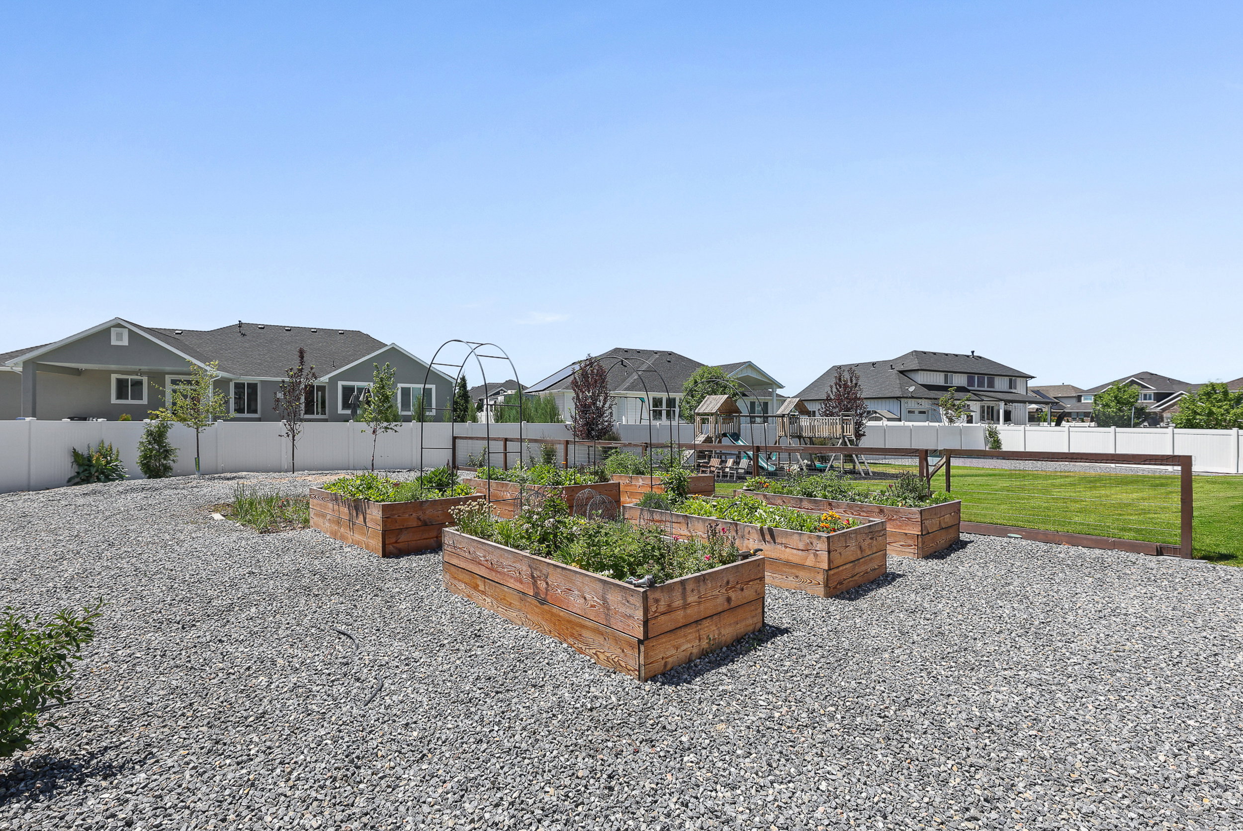 Backyard featuring multiple raised garden beds, gravel ground cover, a white privacy fence, and a section of manicured lawn
