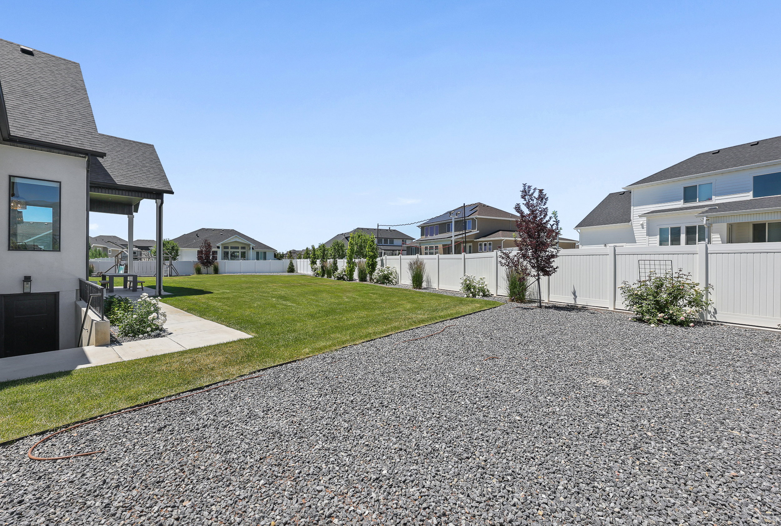Spacious backyard featuring a large gravel area, extensive manicured lawn, white privacy fencing, and a covered patio with dark columns