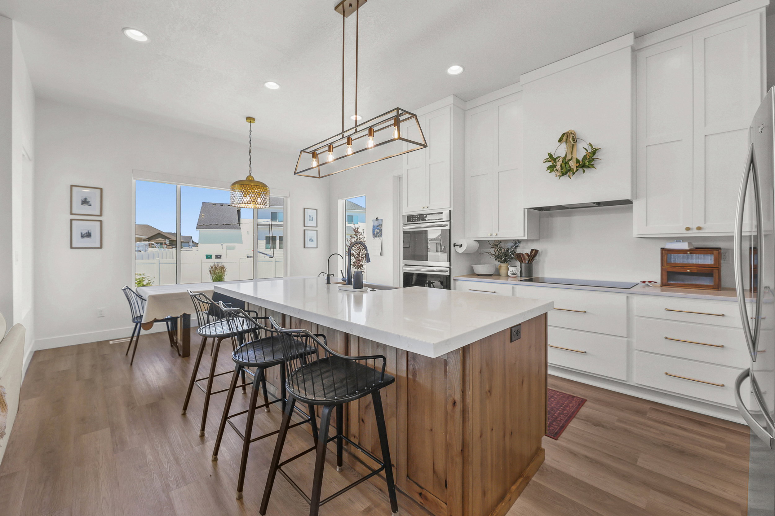 Bright kitchen featuring a substantial island with a light-toned countertop, wood-finish flooring, white cabinetry with gold-toned hardware, stainless steel double ovens, and recessed lighting