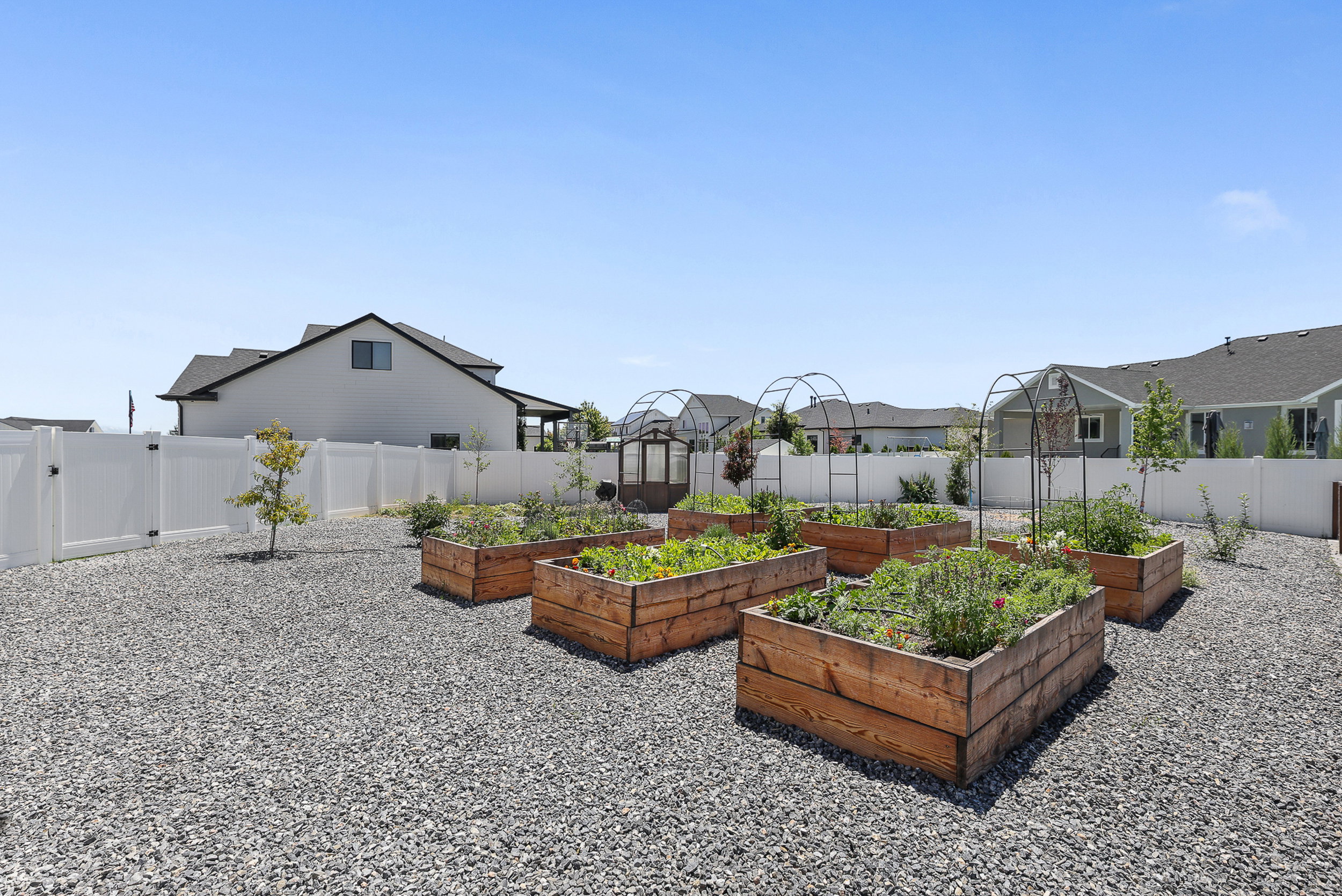 Backyard area featuring multiple raised wooden garden beds with plantings, metal arch trellises, and a gravel ground cover