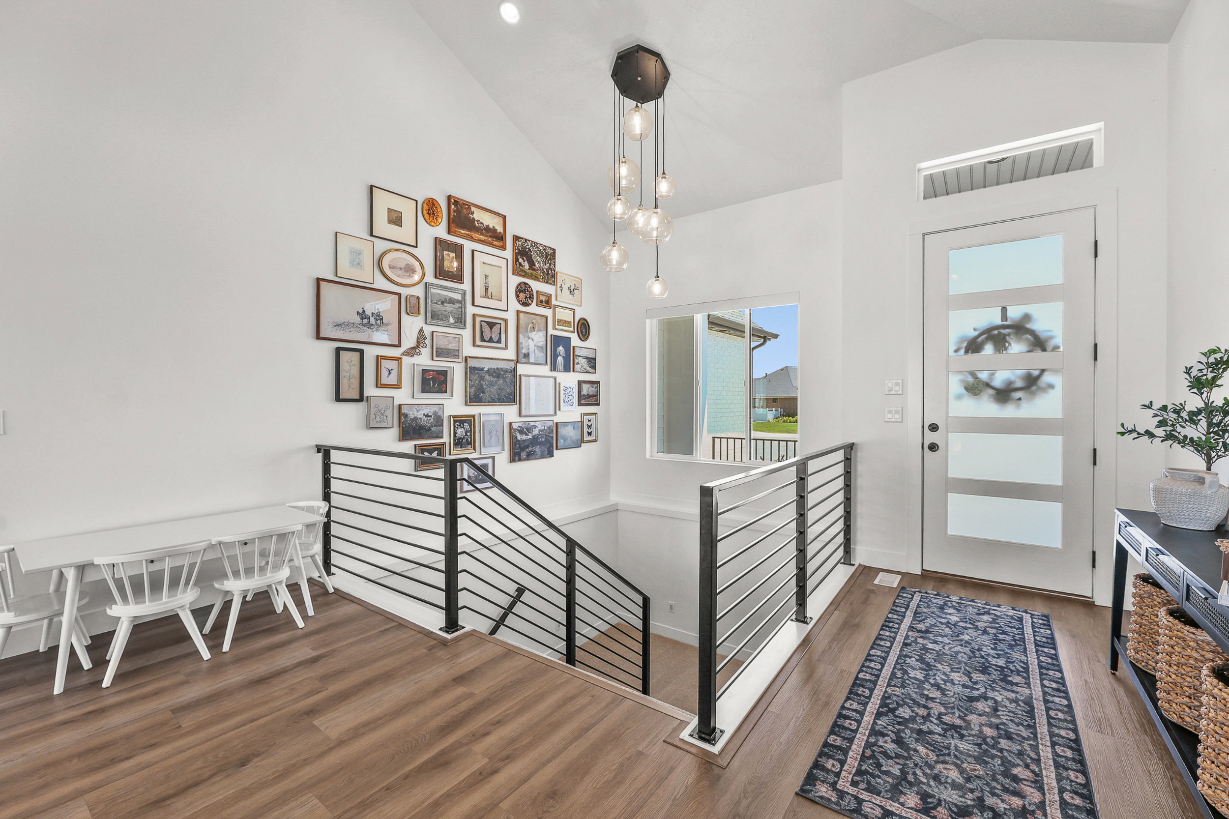 Bright interior entry featuring a contemporary front door with frosted glass panels, wood-finish flooring, a modern multi-pendant light fixture, and sleek metal stair railing
