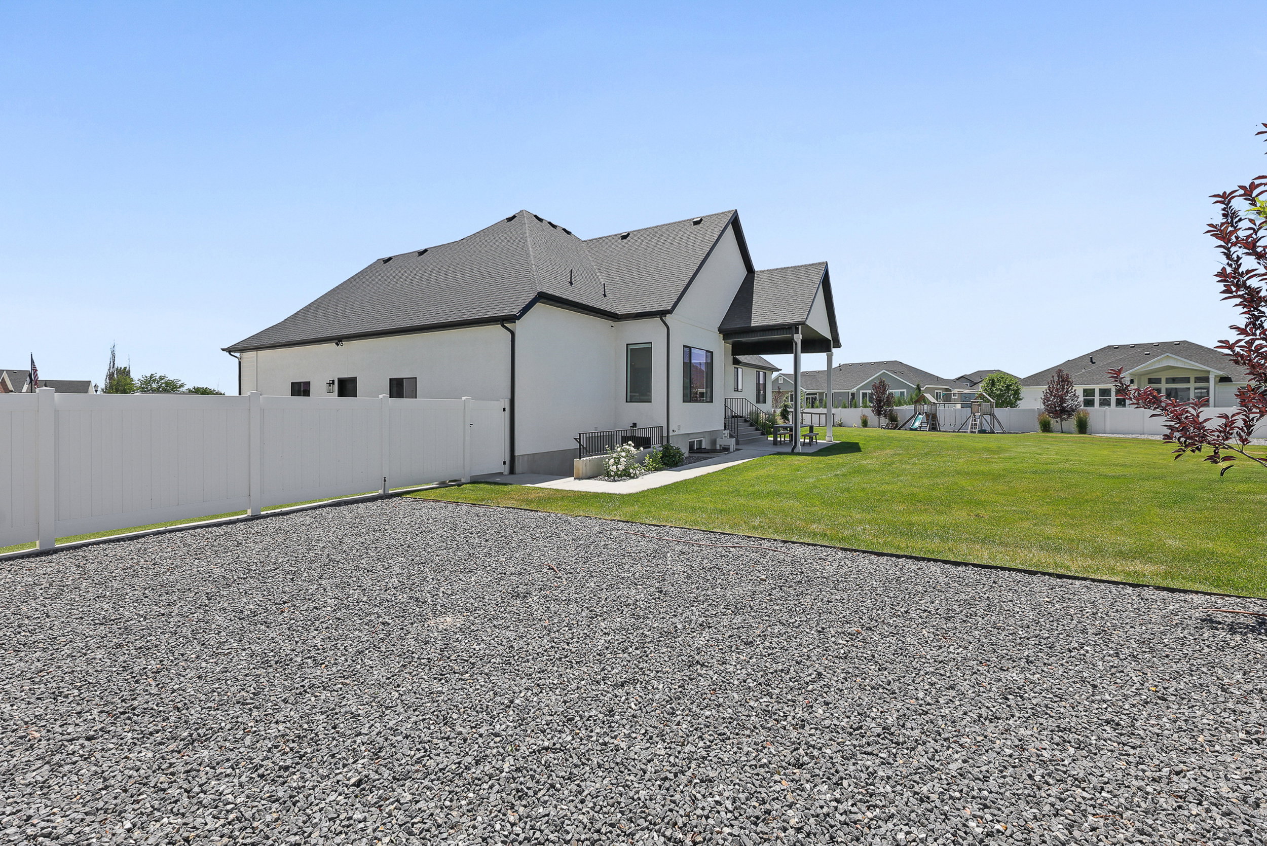 Expansive gravel area bordering a manicured lawn, featuring a white vinyl privacy fence and a covered patio area