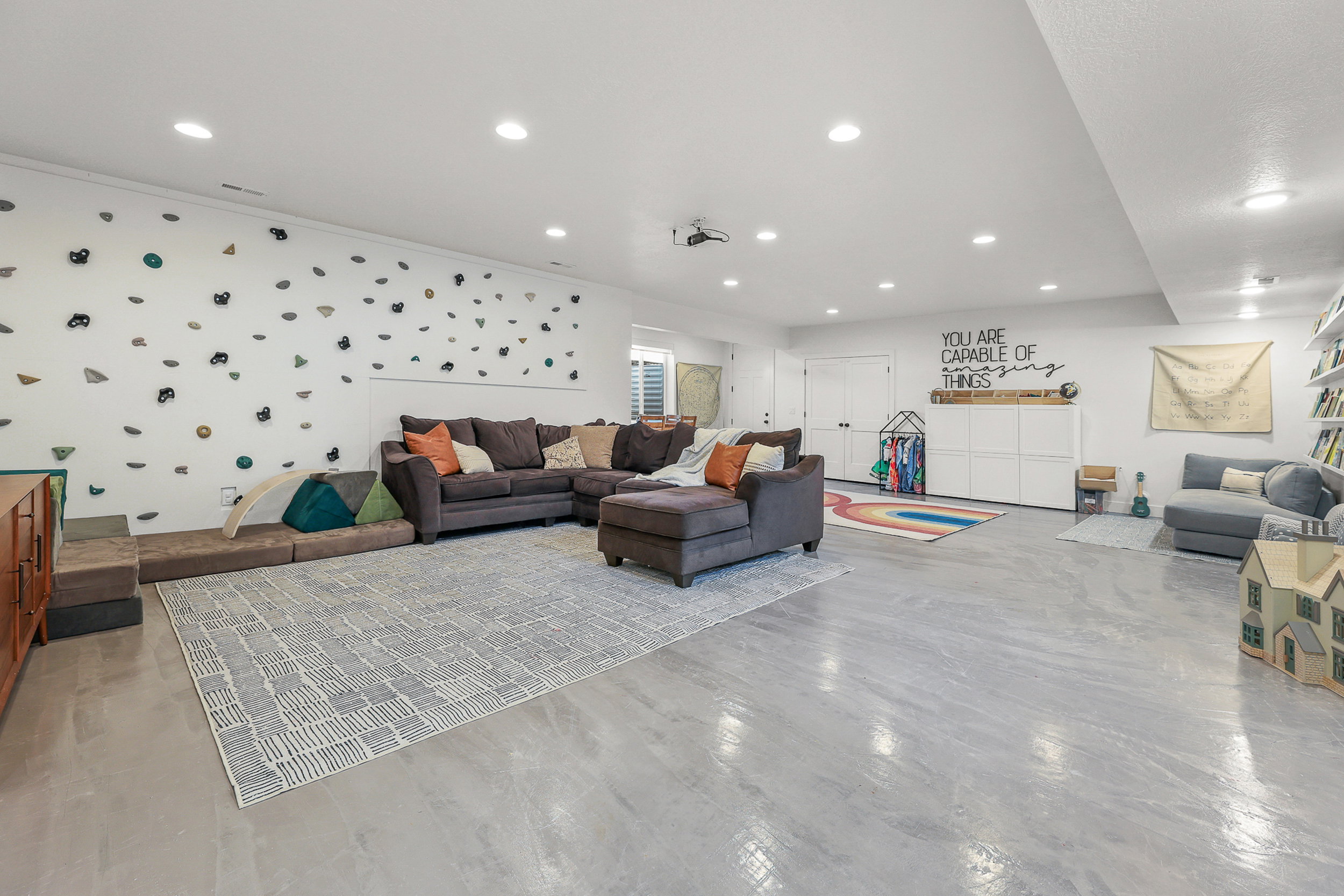 Spacious recreation room featuring light gray flooring, recessed lighting, and a white accent wall with climbing holds