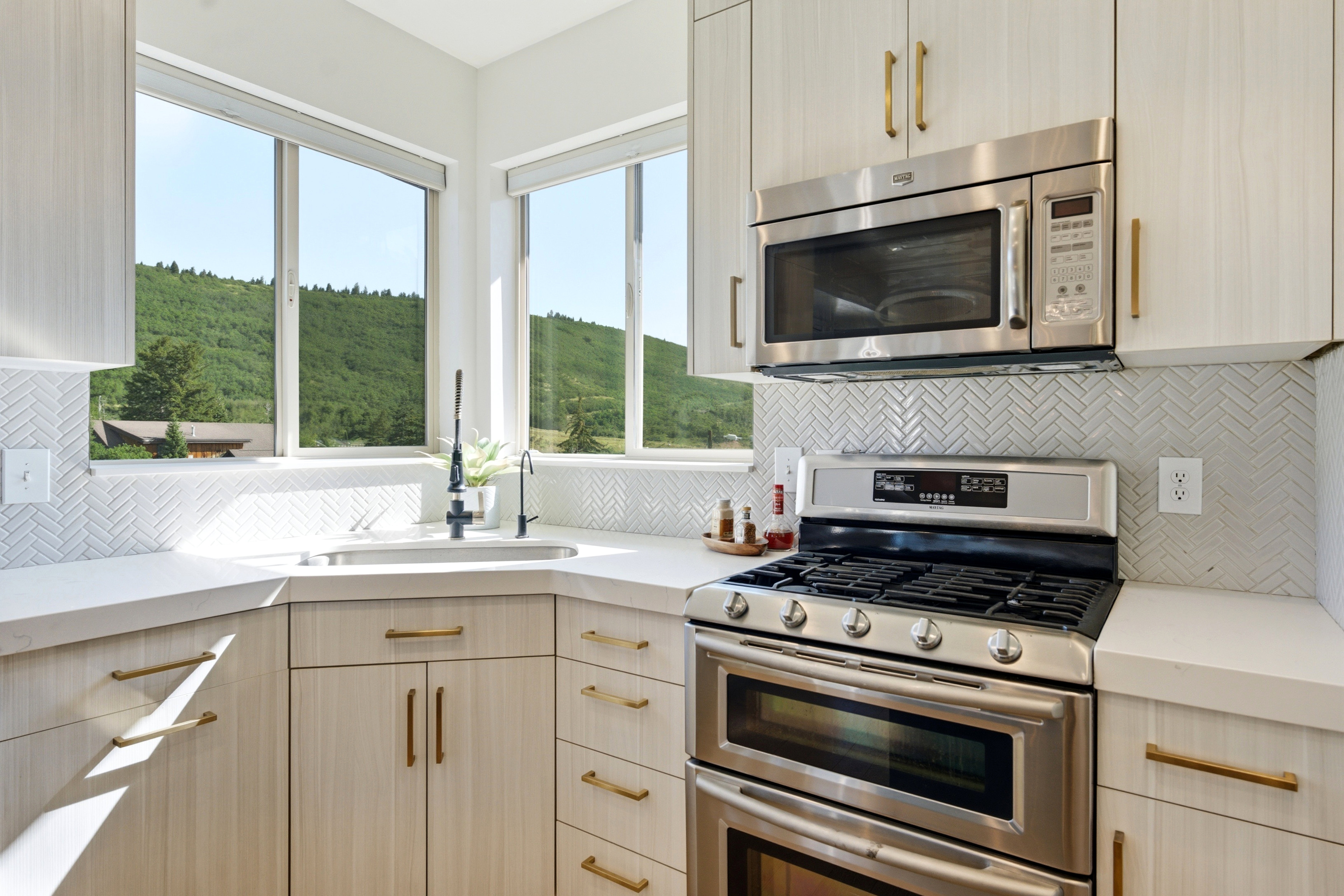 Contemporary kitchen featuring light wood-finish cabinetry with brass hardware, stainless steel appliances, a corner sink, and white herringbone tile backsplash