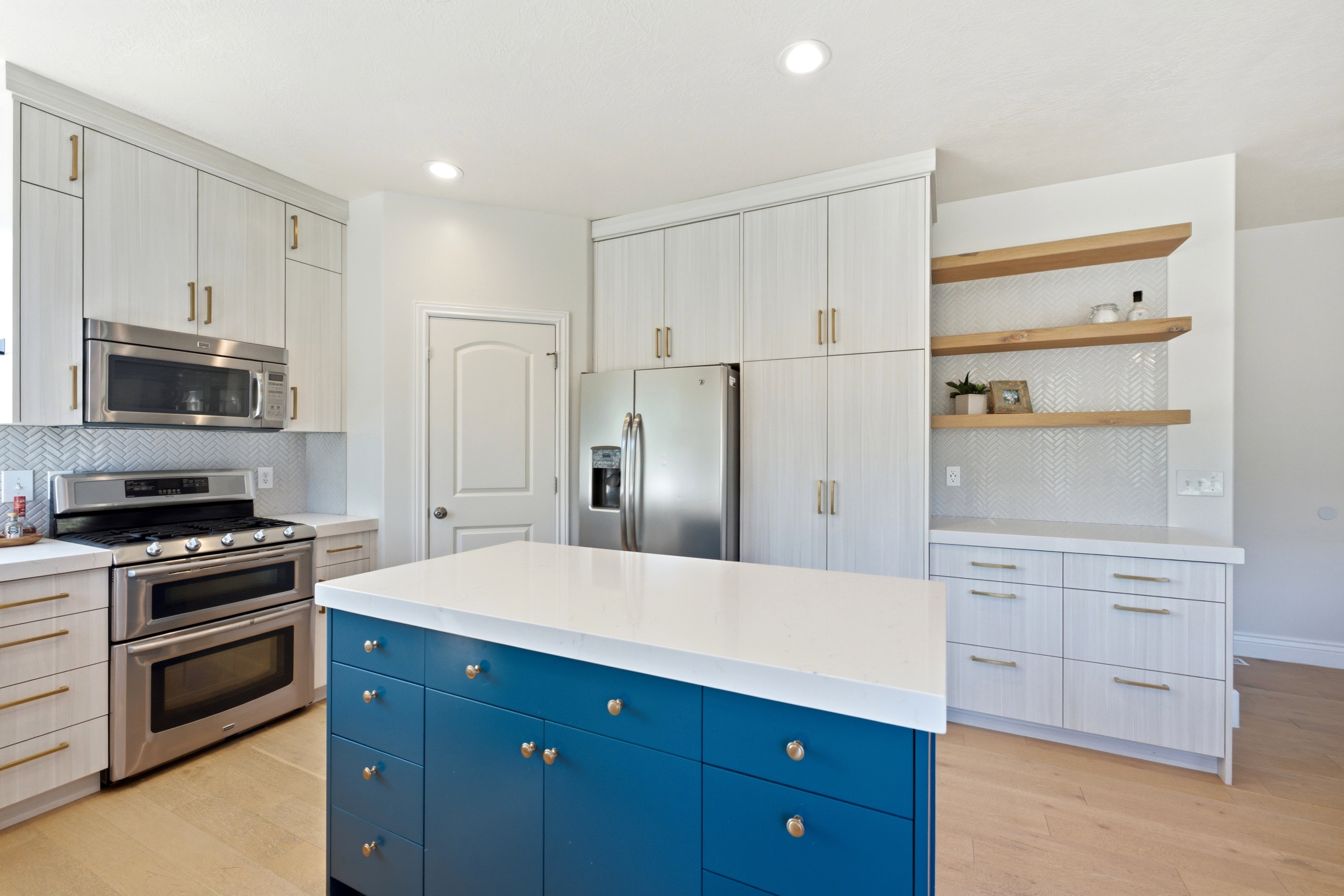 Gourmet kitchen featuring a vibrant blue island with a white countertop, stainless steel appliances, light wood-finish cabinetry with gold-tone hardware, a white herringbone tile backsplash, and light wood-finish flooring