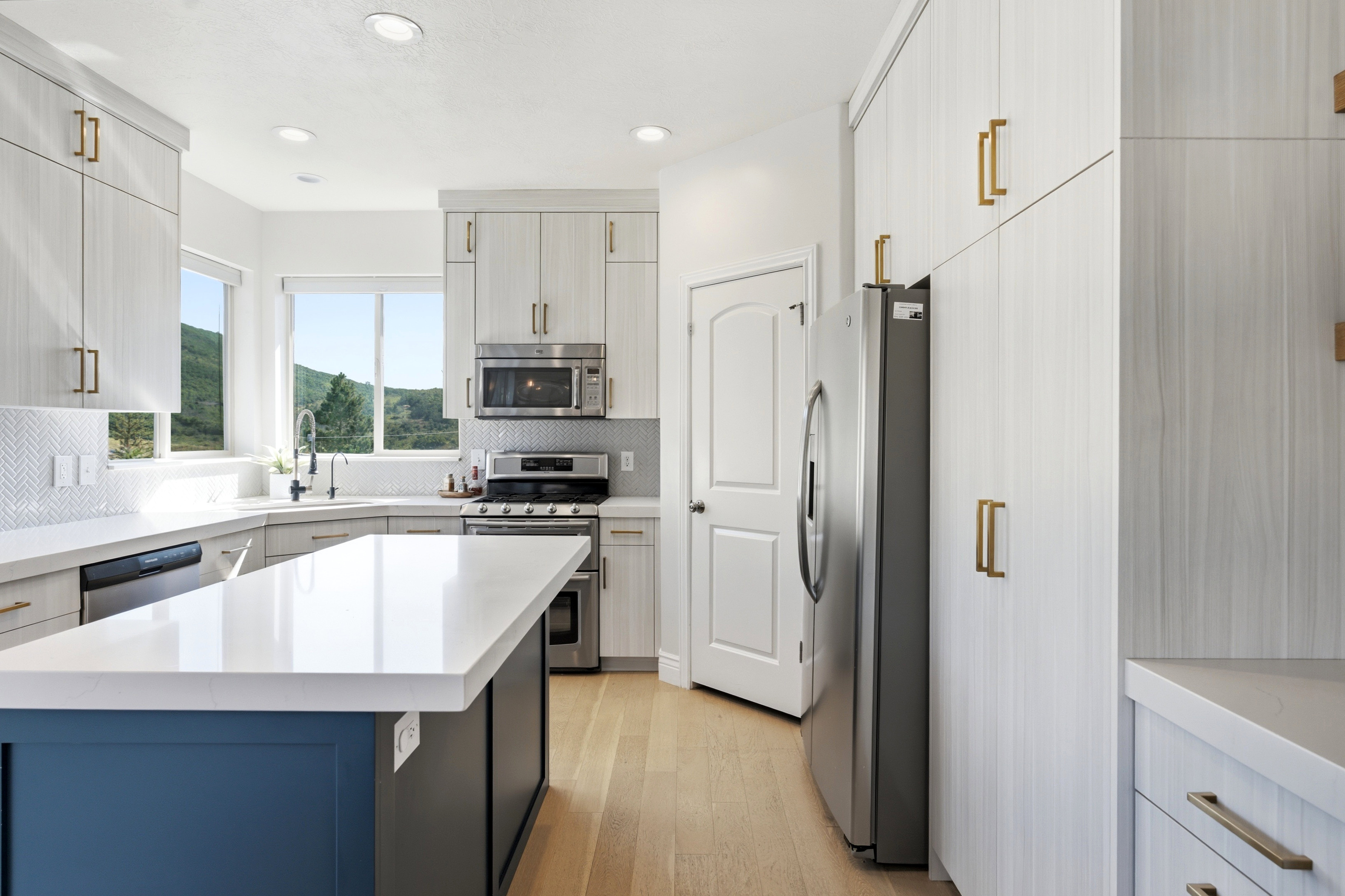 Modern kitchen featuring a central island with a contrasting blue base, light wood-finish flooring, white countertops, built-in stainless steel appliances, and abundant light-toned cabinetry with gold-toned hardware