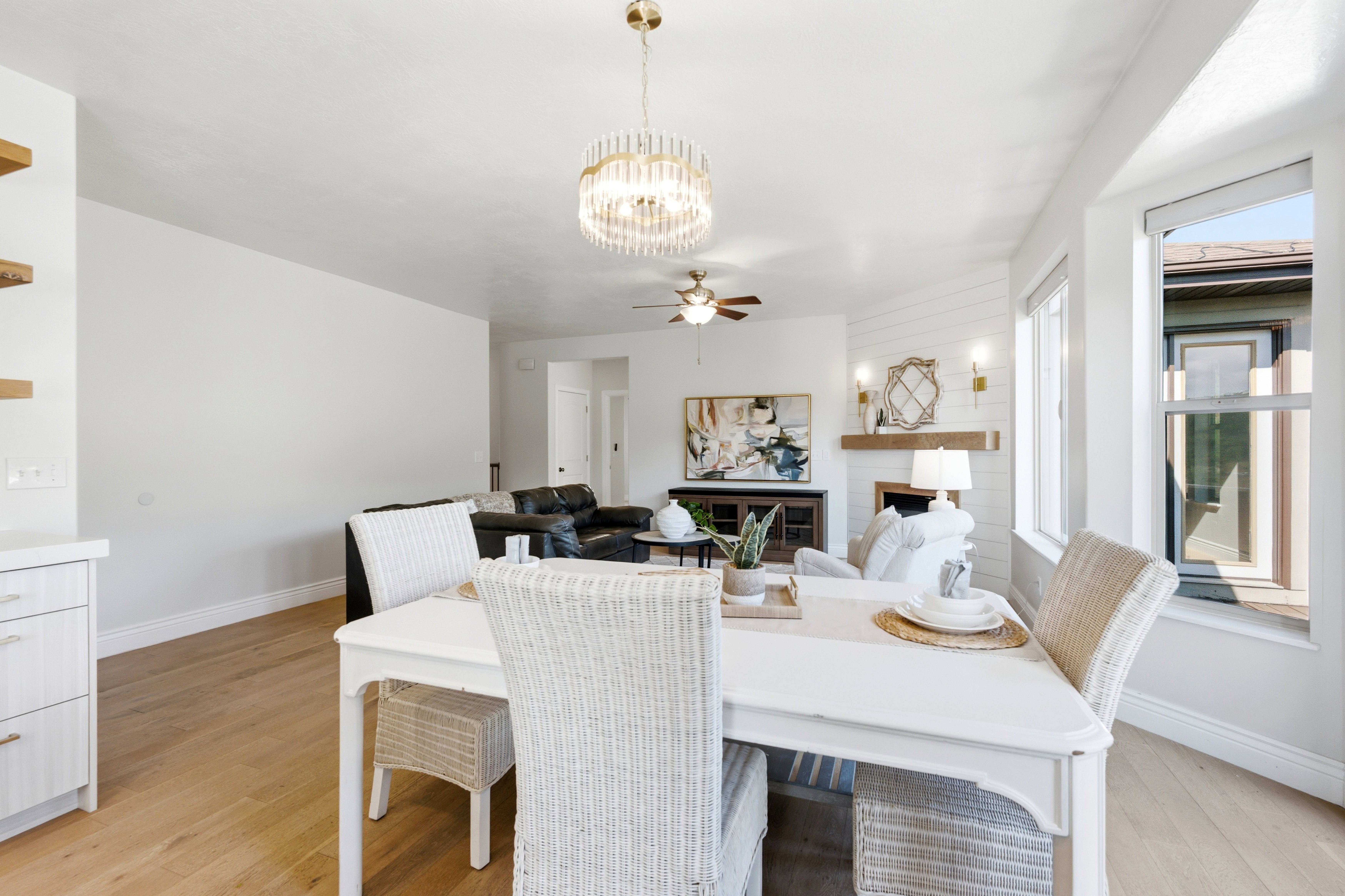 Open-concept interior featuring light wood-finish flooring, a contemporary chandelier, a white fireplace with a natural wood mantel, shiplap accent wall, and multiple windows