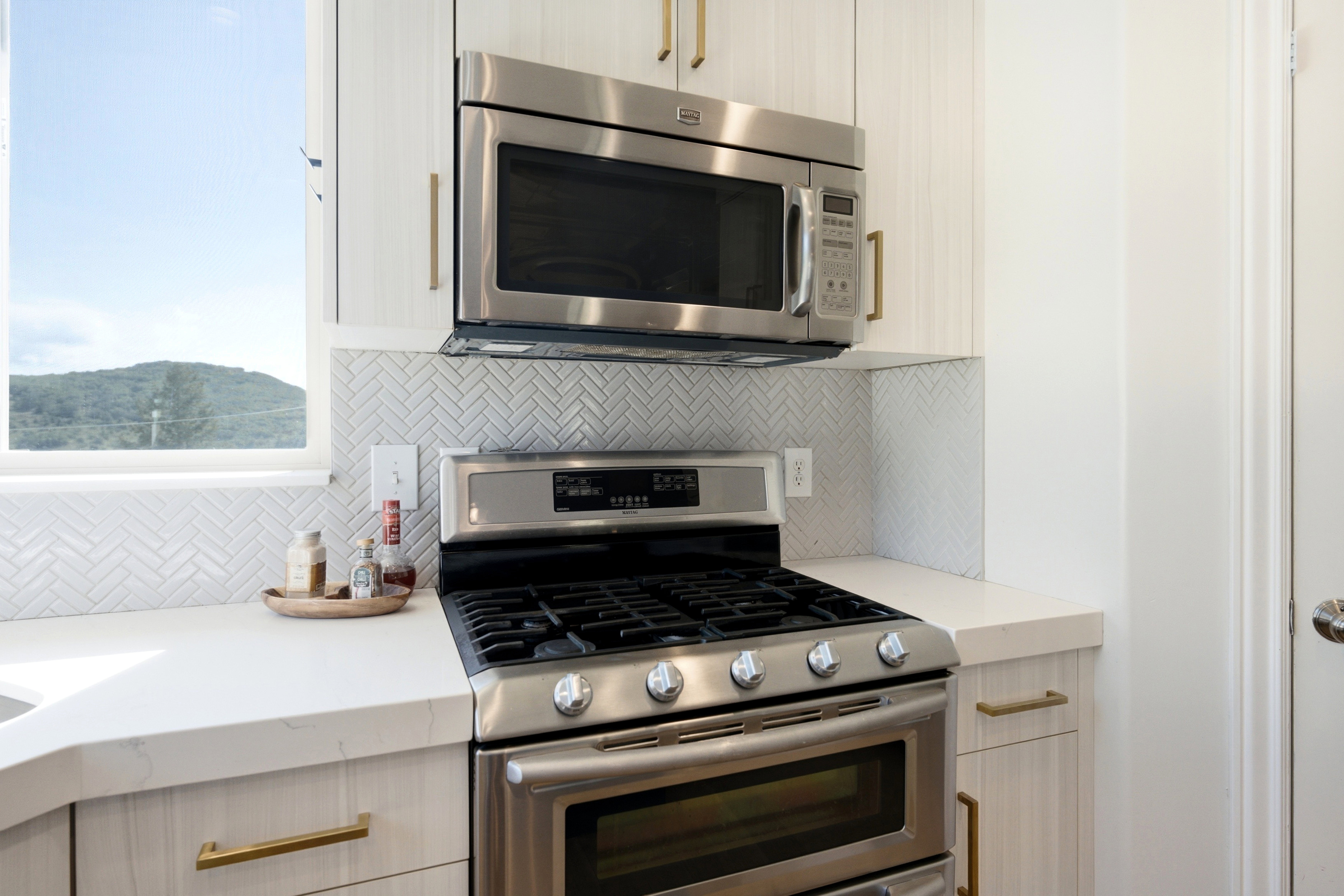 Kitchen featuring stainless steel gas range, coordinating microwave, white herringbone tile backsplash, light wood-finish cabinetry, and white countertops